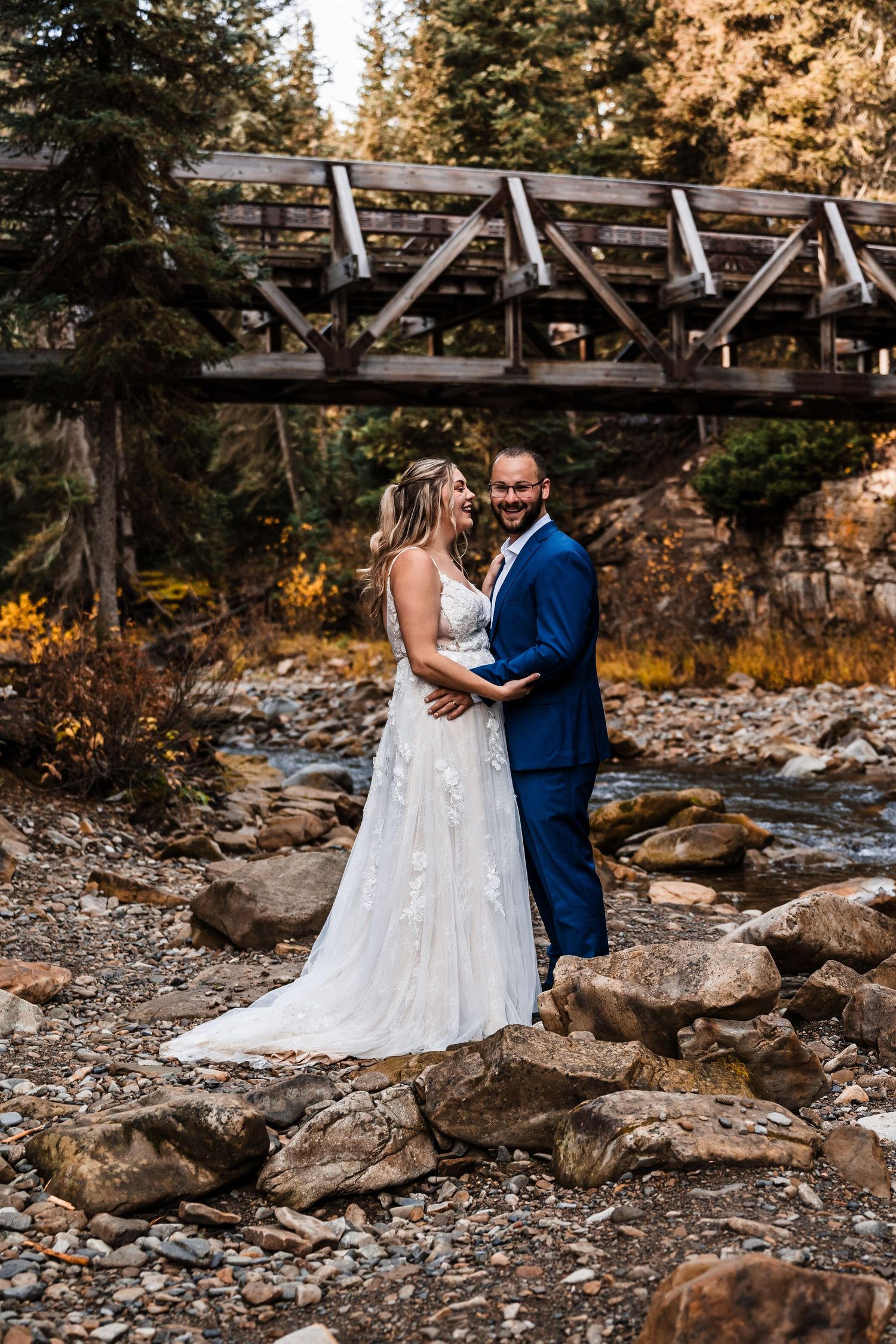 A couple poses on rocky terrain near a wooden bridge during their wedding photoshoot.