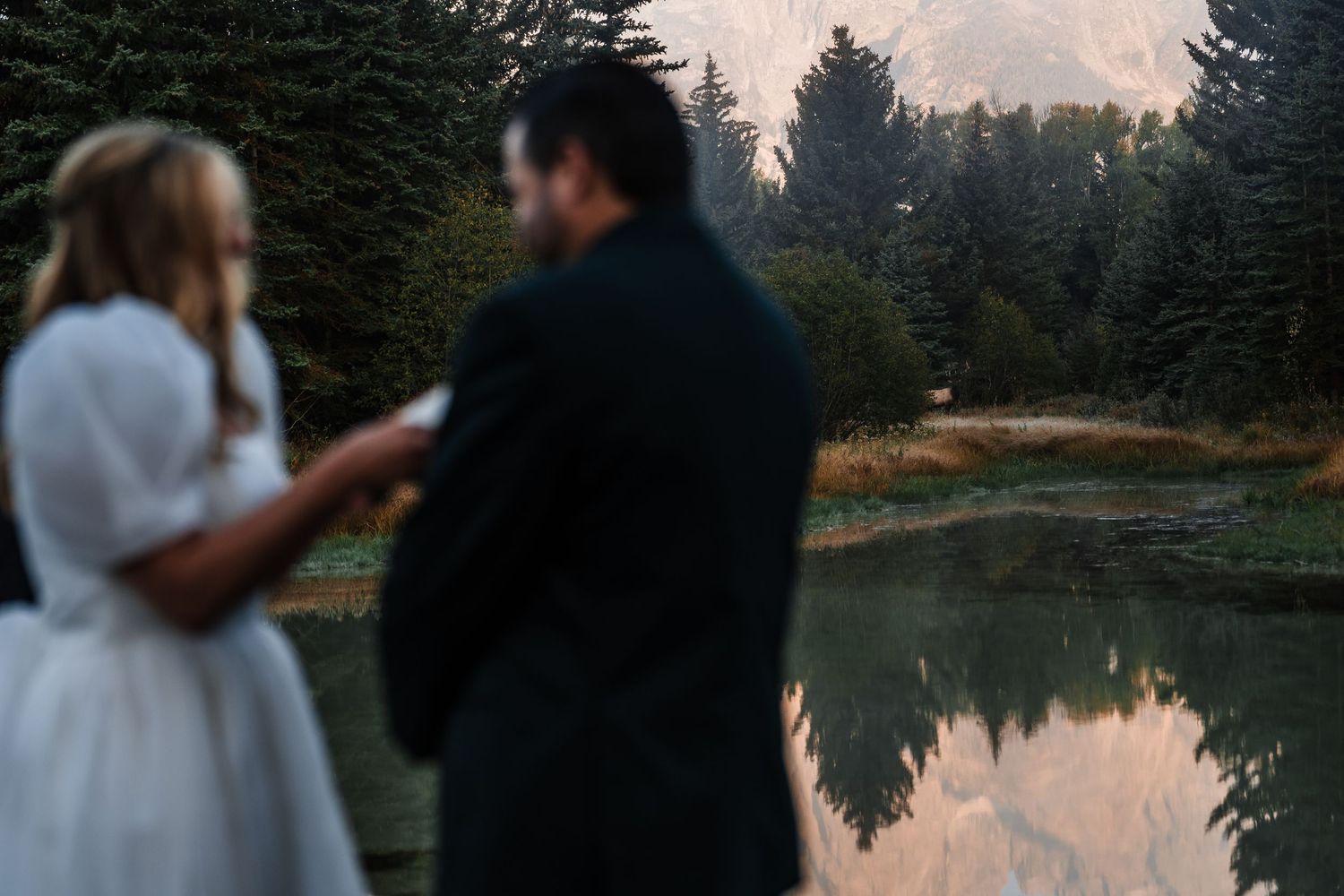 Couple sharing a romantic moment by a serene mountain lake during sunset with evergreen trees in the background.