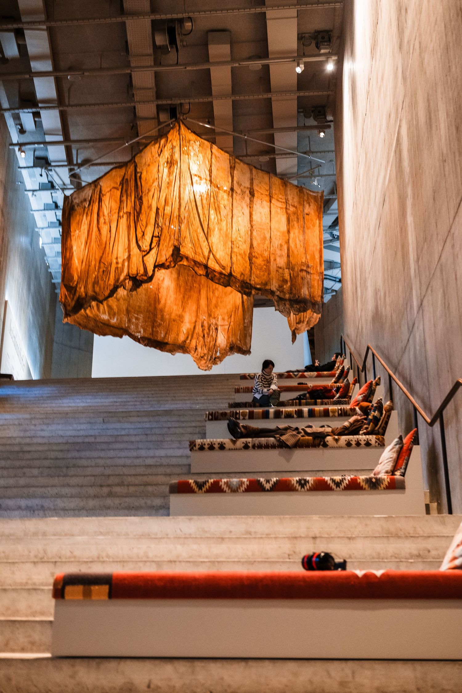 Art installation features large orange fabric suspended from ceiling above steps in gallery space.
