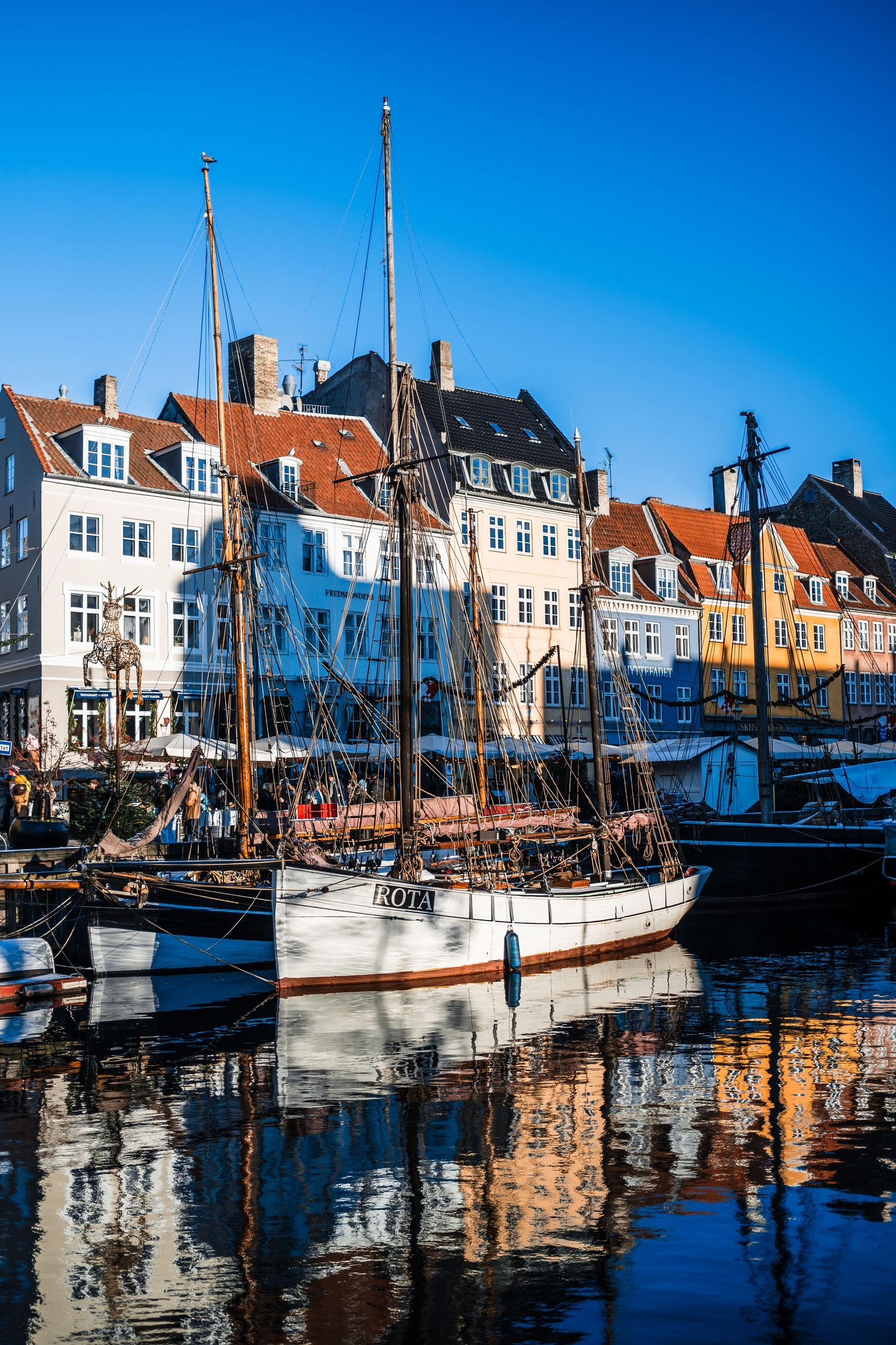 Historic wooden sailing ships docked along colorful buildings in Nyhavn canal harbor of Copenhagen on a sunny day.