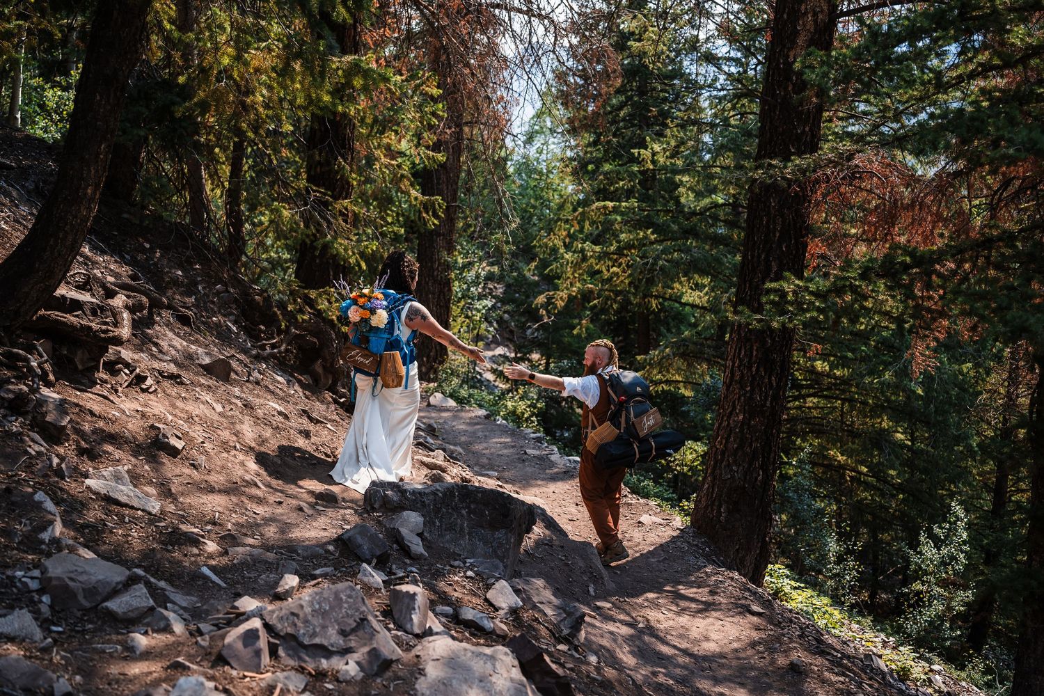 Bride and groom hiking through sunlit forest trail in wedding attire carrying backpacks.