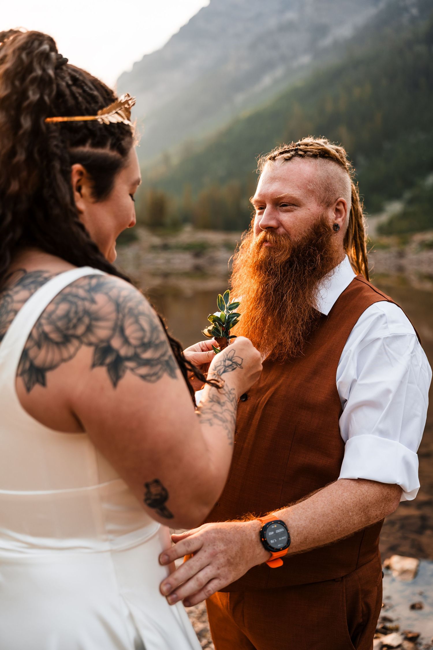Groom with red beard and dreadlocks smiles as tattooed bride pins boutonniere on brown vest at mountain elopement.