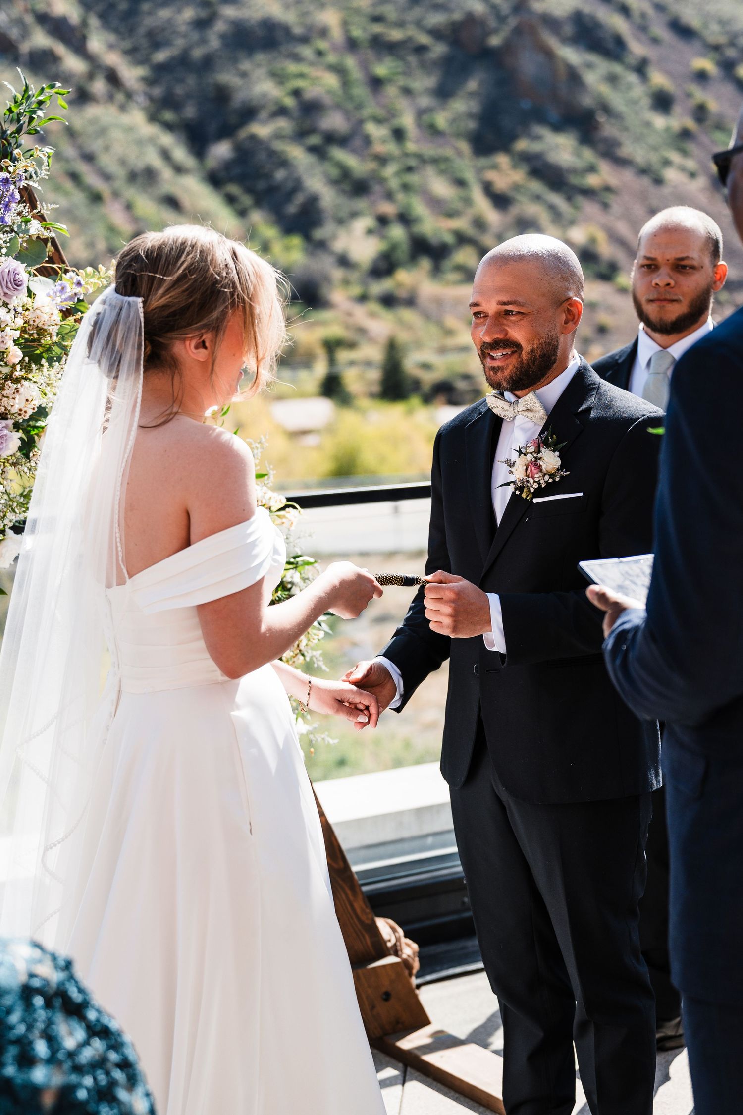 Groom smiling at bride during outdoor wedding ceremony with mountain backdrop and floral arch.