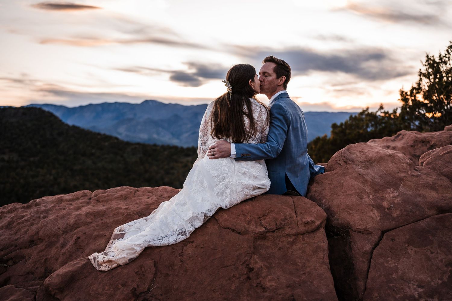 Bride and groom sharing a kiss on red rocks with mountain sunset view behind them.