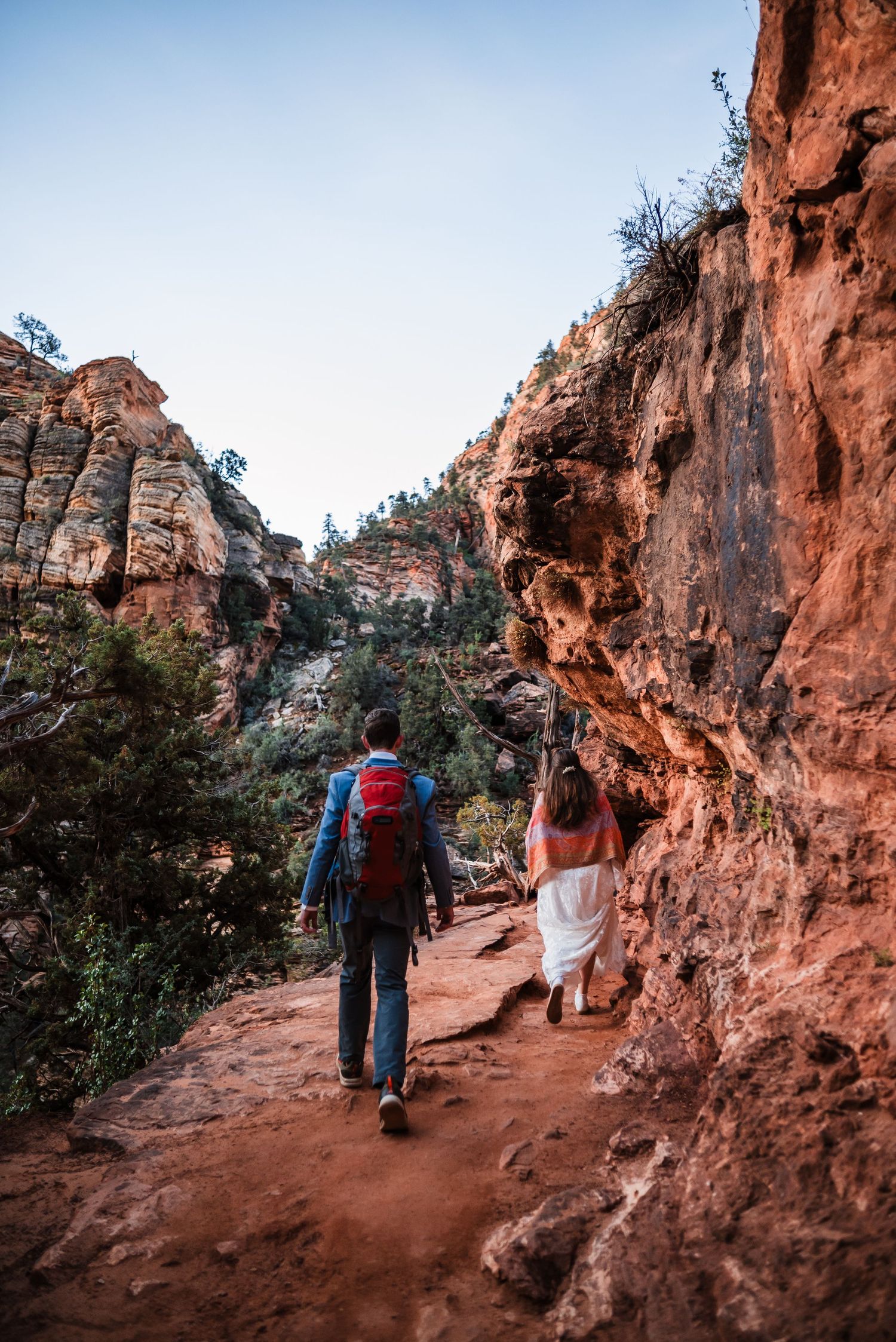 Couple hiking through red rock canyon trail in Zion National Park, bride in white dress and groom with red backpack.