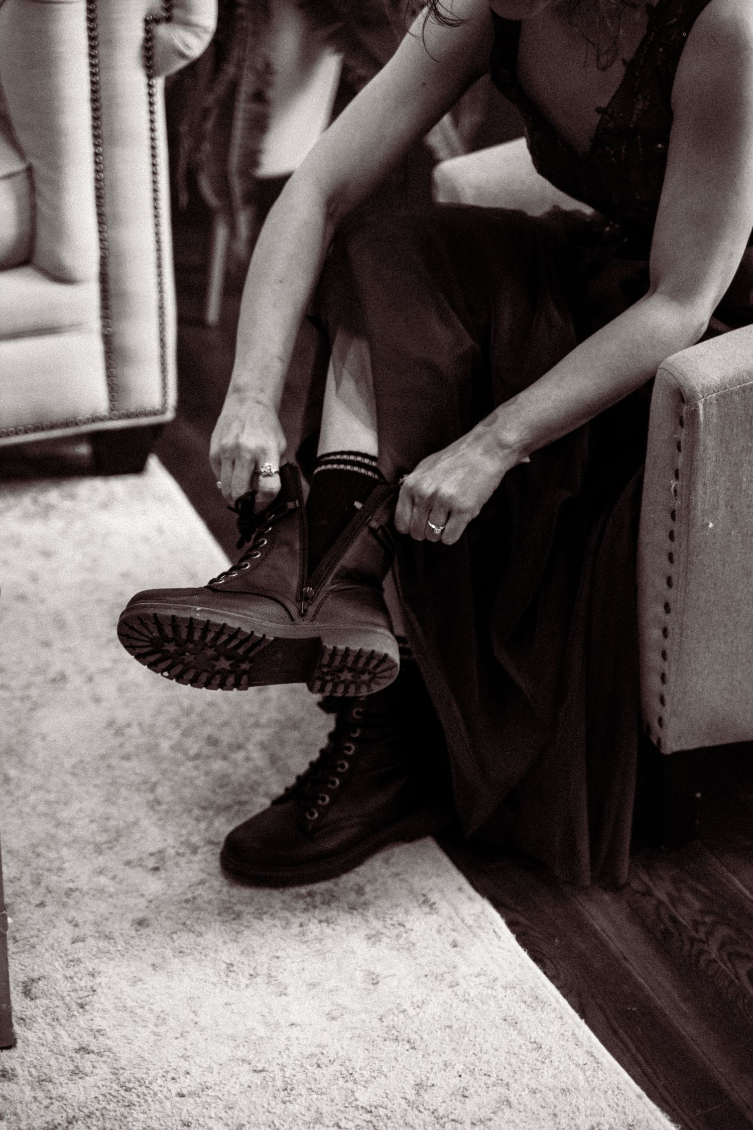 Black and white photo of a bride lacing up combat boots under her dark wedding gown while seated.