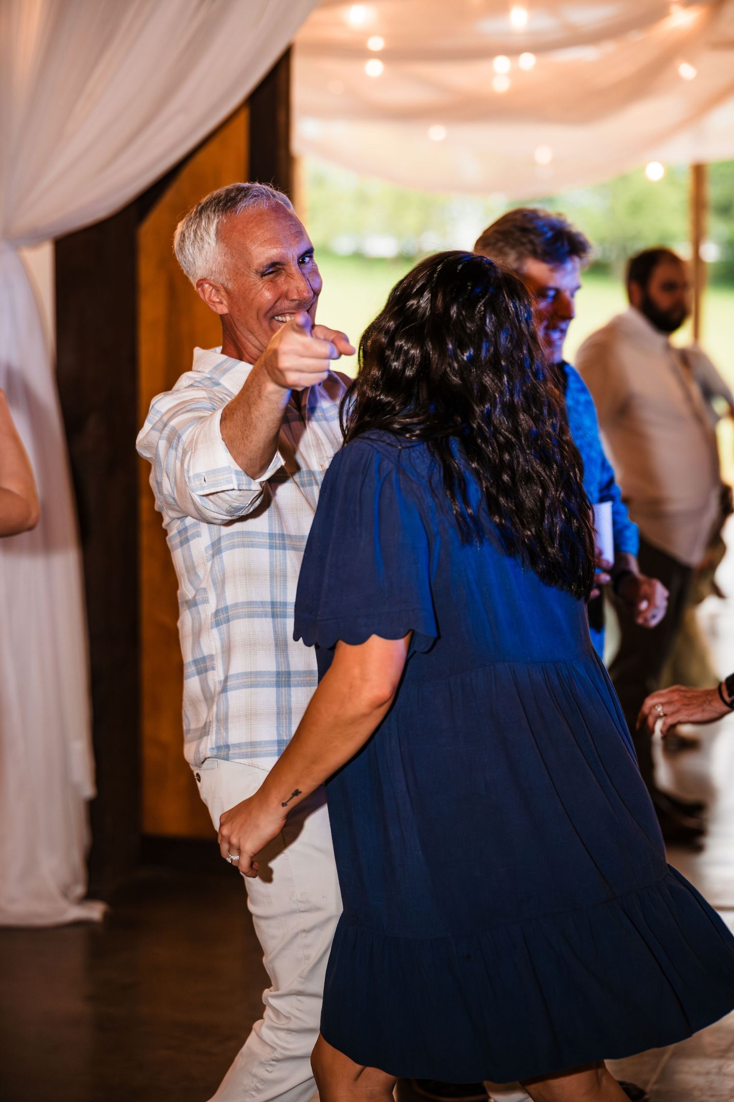 Guests dance joyfully at a wedding reception, with a man in a plaid shirt smiling and pointing.