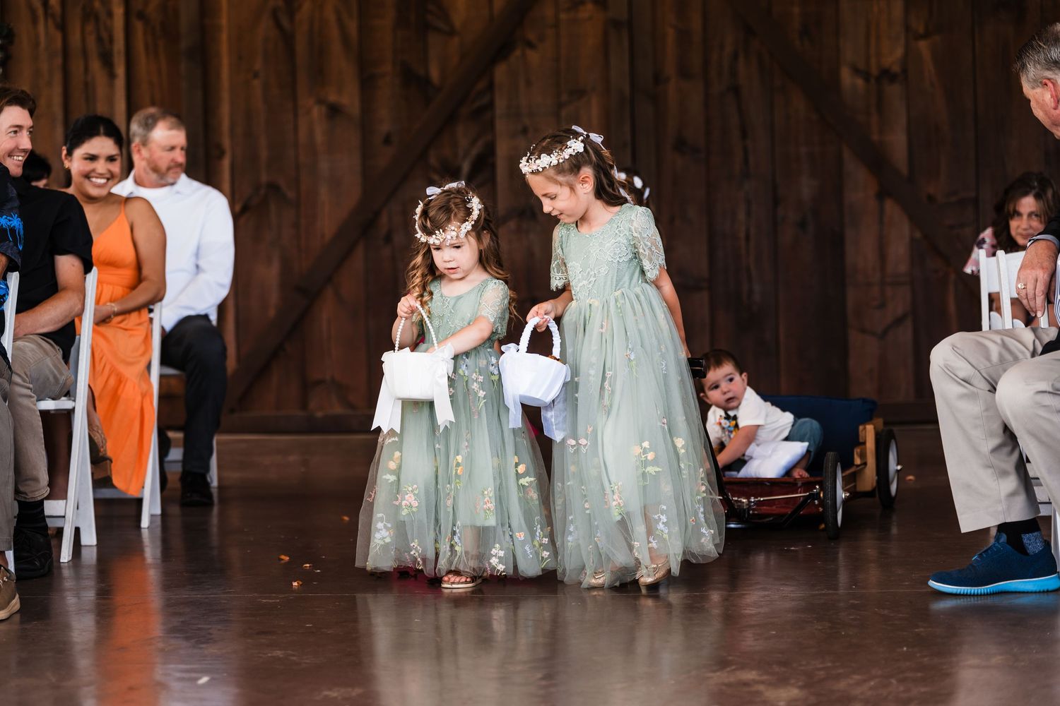 Two flower girls in sage green dresses walk down barn wedding aisle.