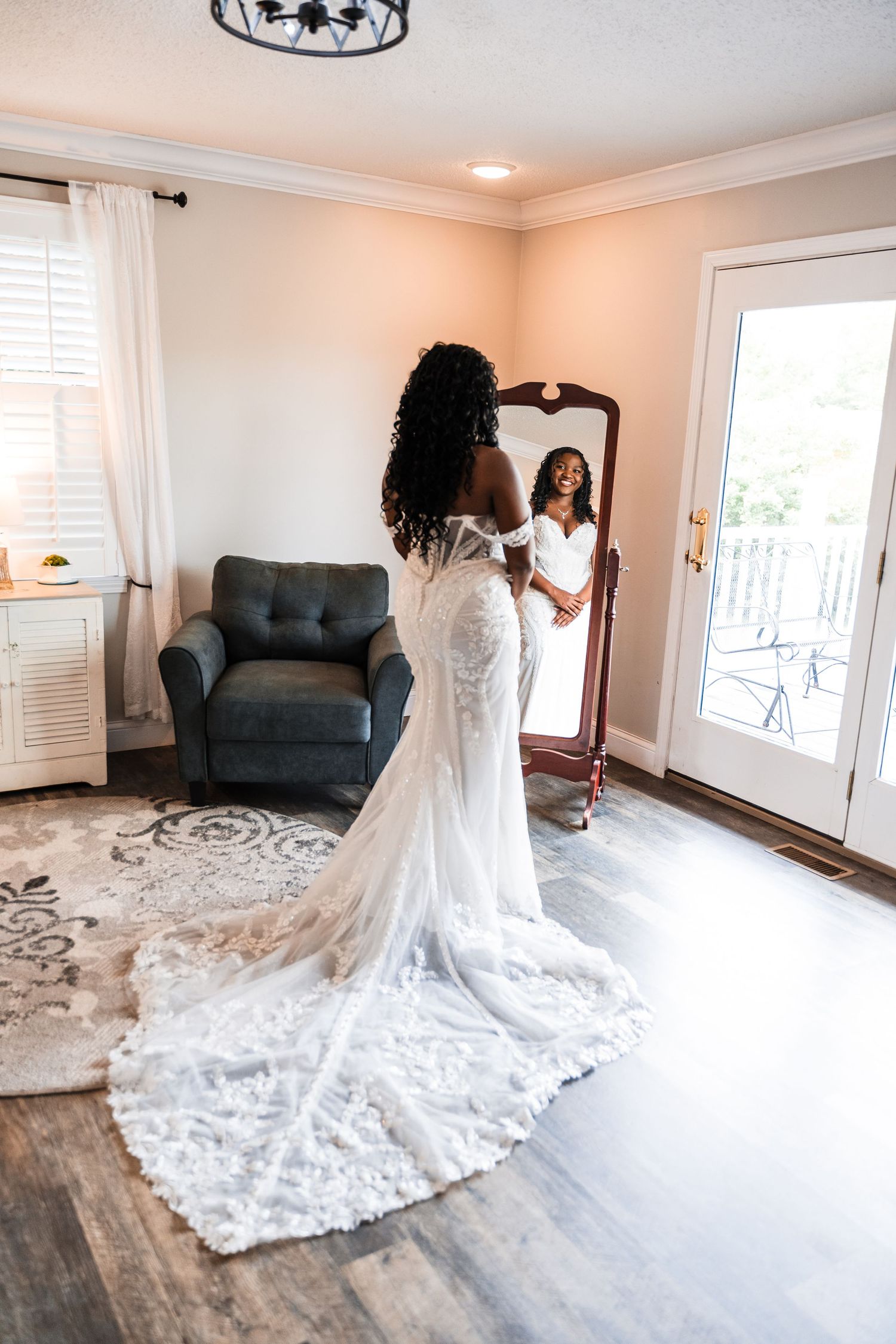 Bride in lace wedding gown with train admires her reflection in a full-length mirror in a bright elegant room.