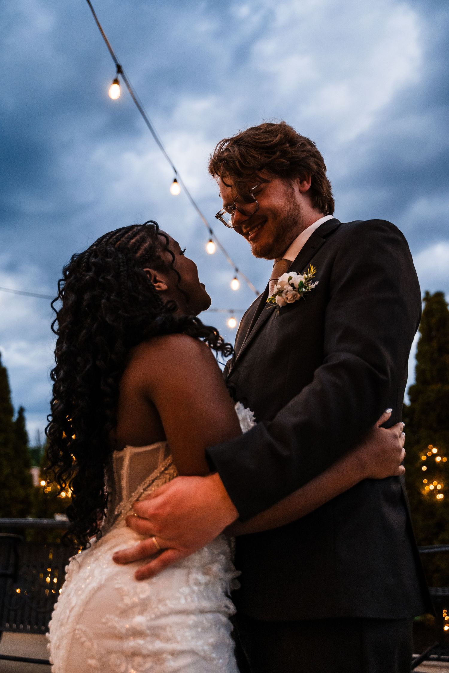Bride and groom embracing under string lights at dusk with smiling expressions at wedding.