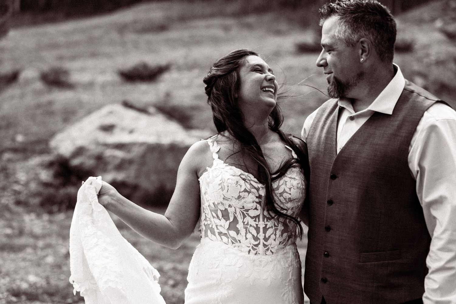 Black and white photo of a laughing bride and groom in a vest sharing a joyful outdoor moment.