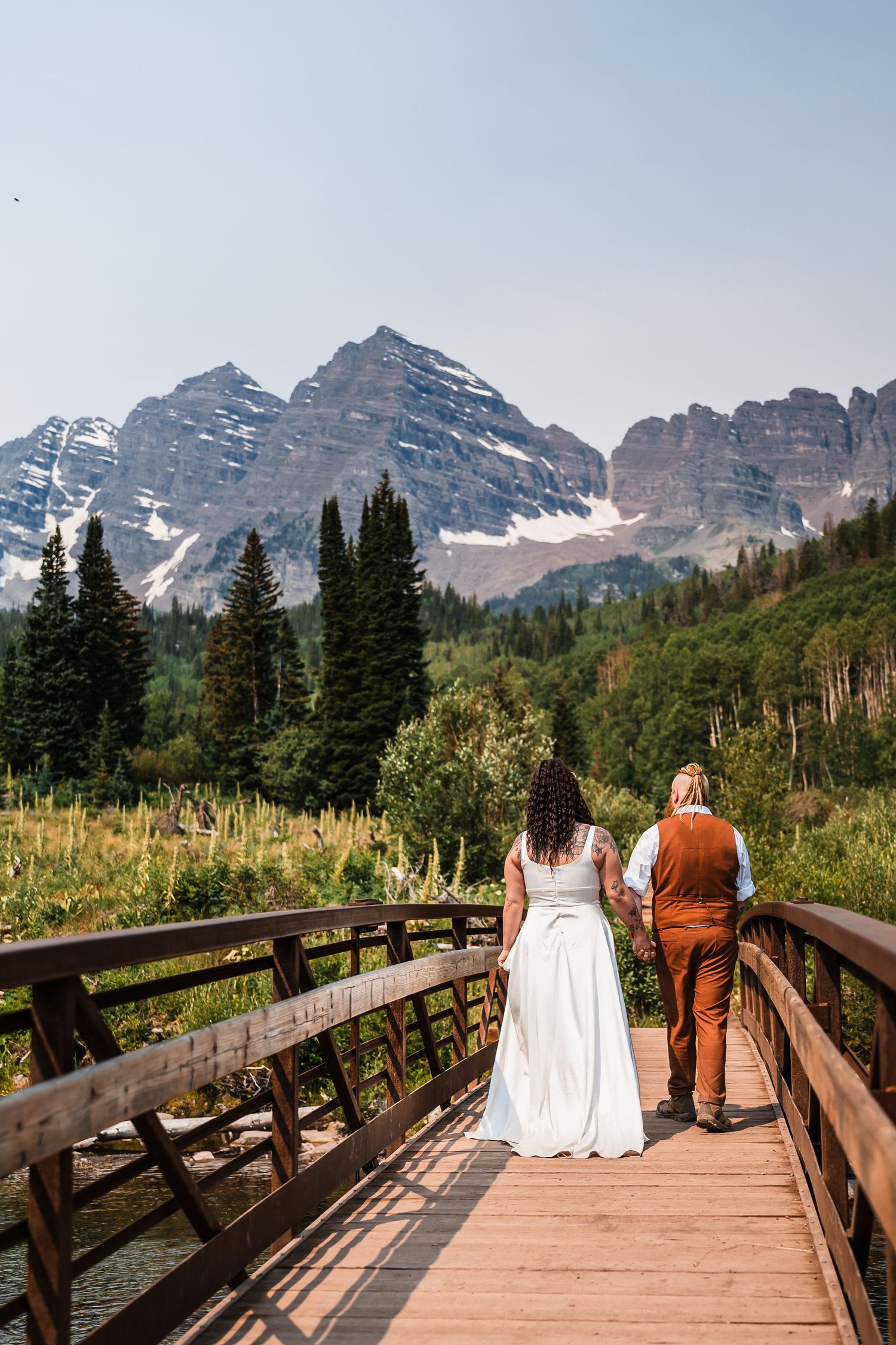 Wedding couple holds hands on wooden bridge overlooking Maroon Bells mountains in Colorado.