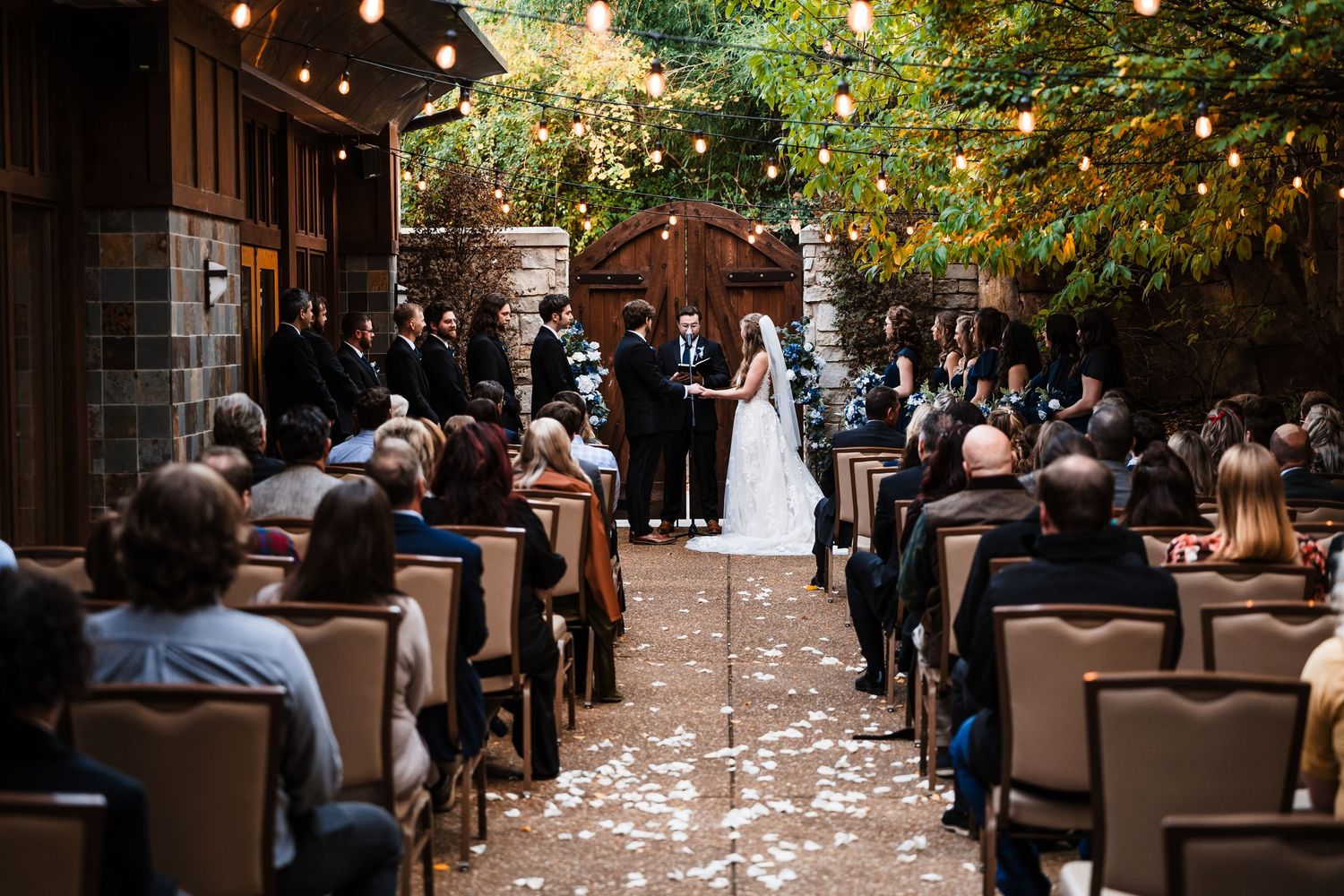 Wedding guests gather in a rustic outdoor courtyard with string lights and scattered flower petals creating a romantic ambiance.