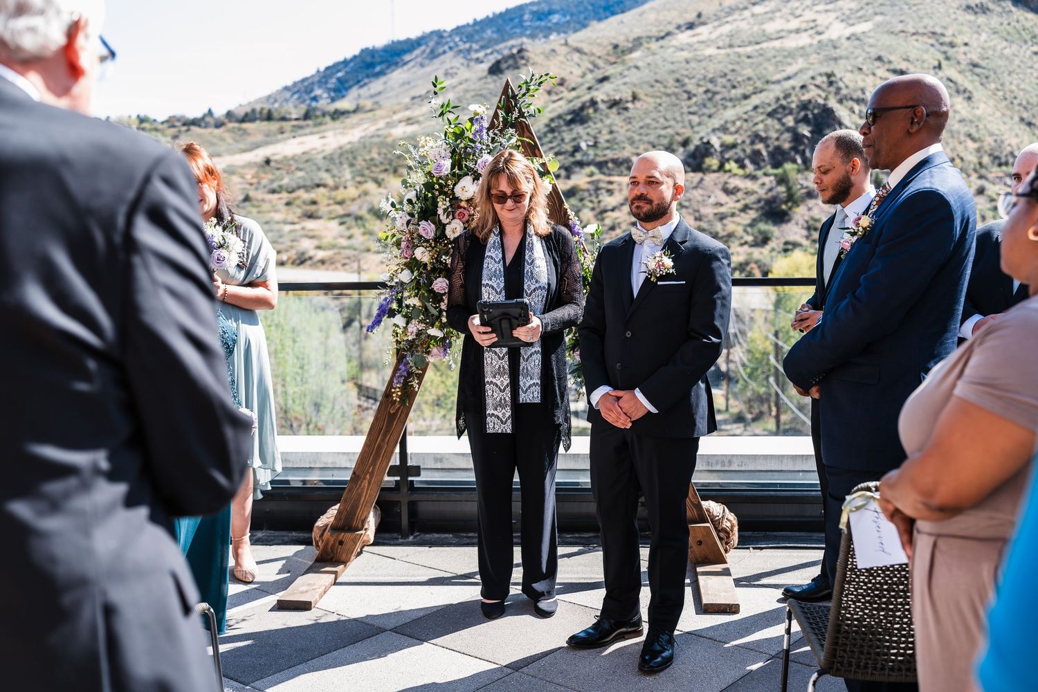 Outdoor rooftop wedding ceremony with officiant and groom standing at floral arch, mountain landscape backdrop.
