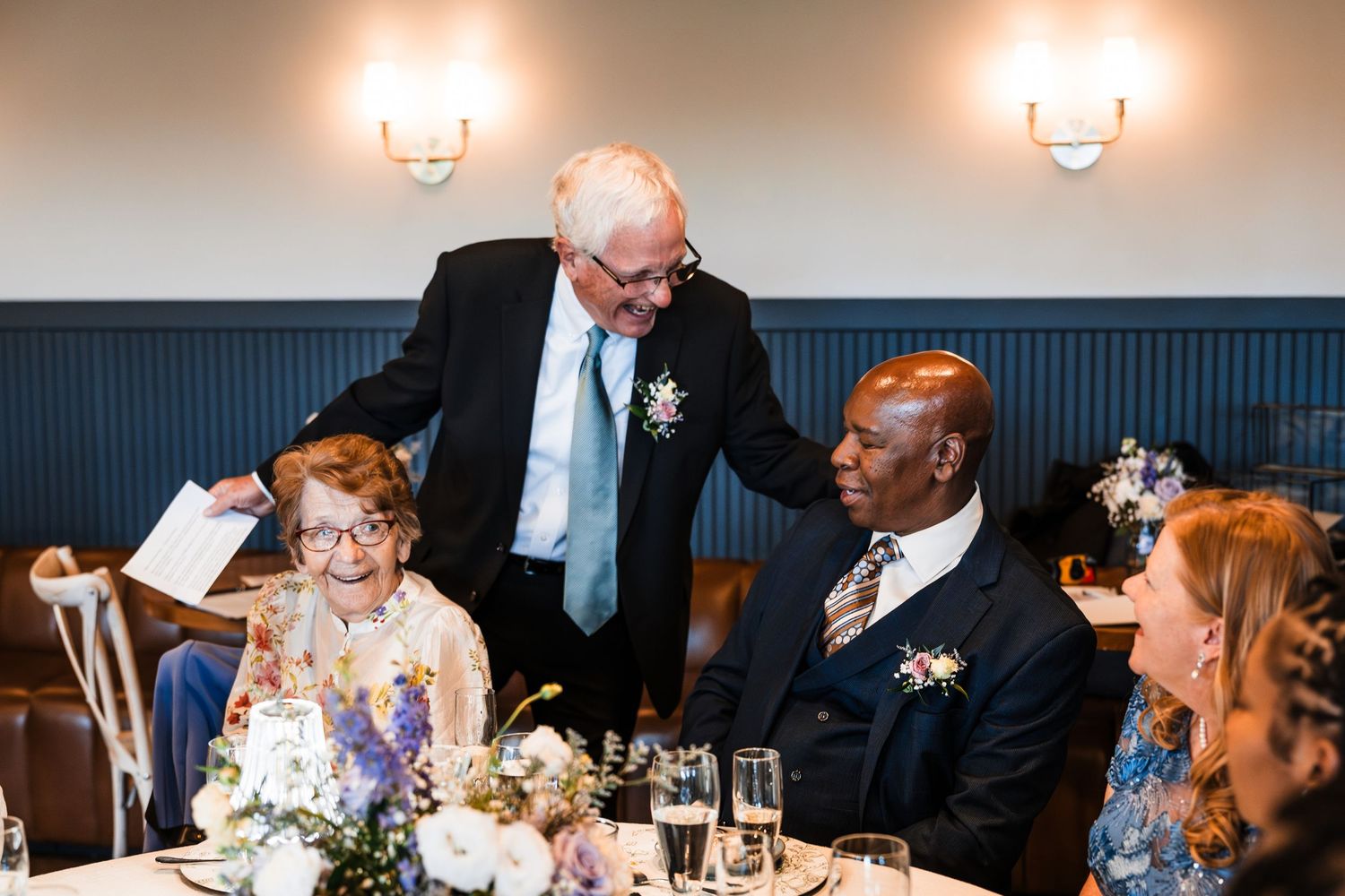 Elderly wedding guest chatting with groom and seated guests at elegant wedding reception dinner.