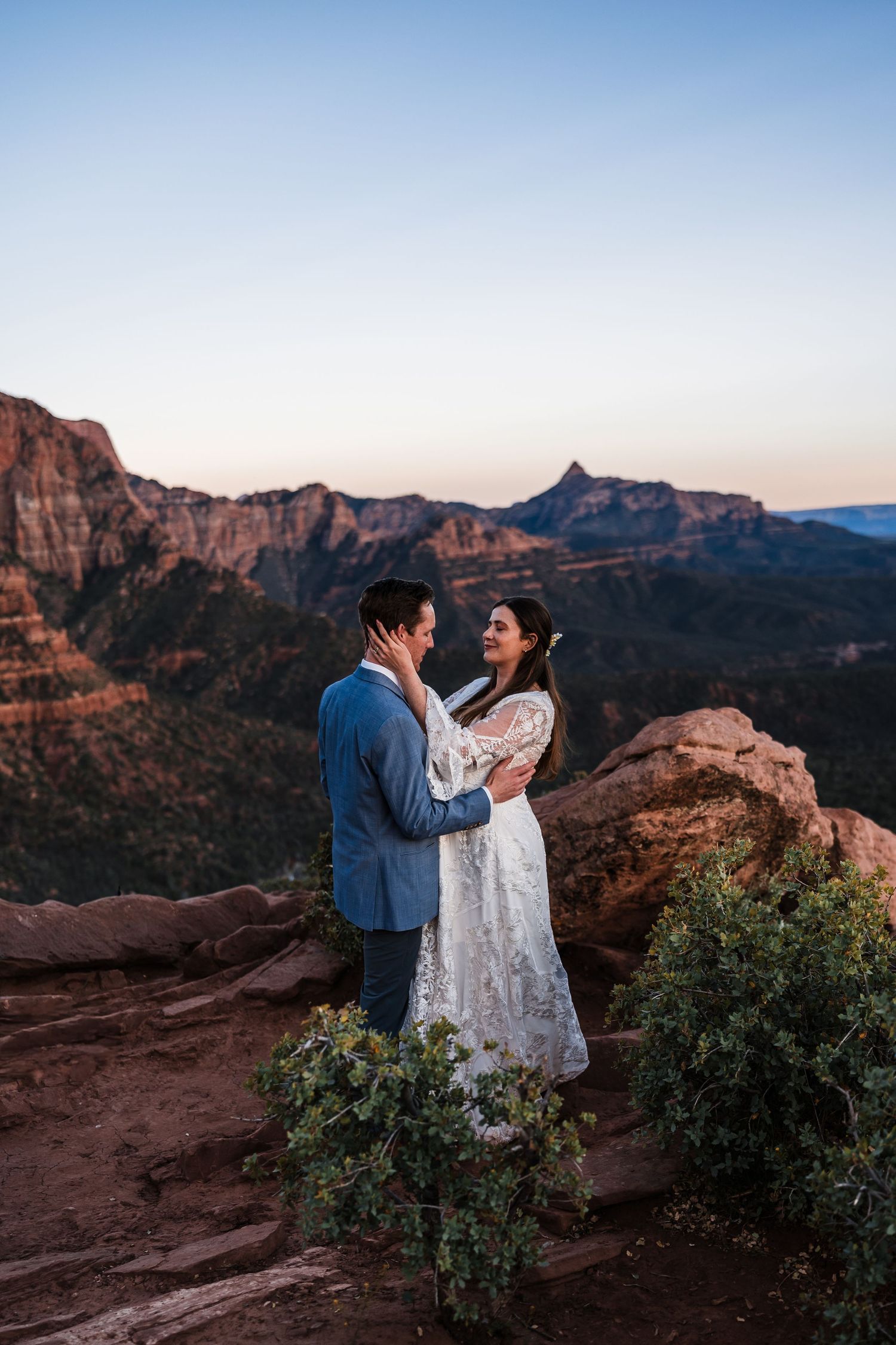 Bride and groom embracing at dusk on a rocky overlook with sweeping Zion National Park mountain views.