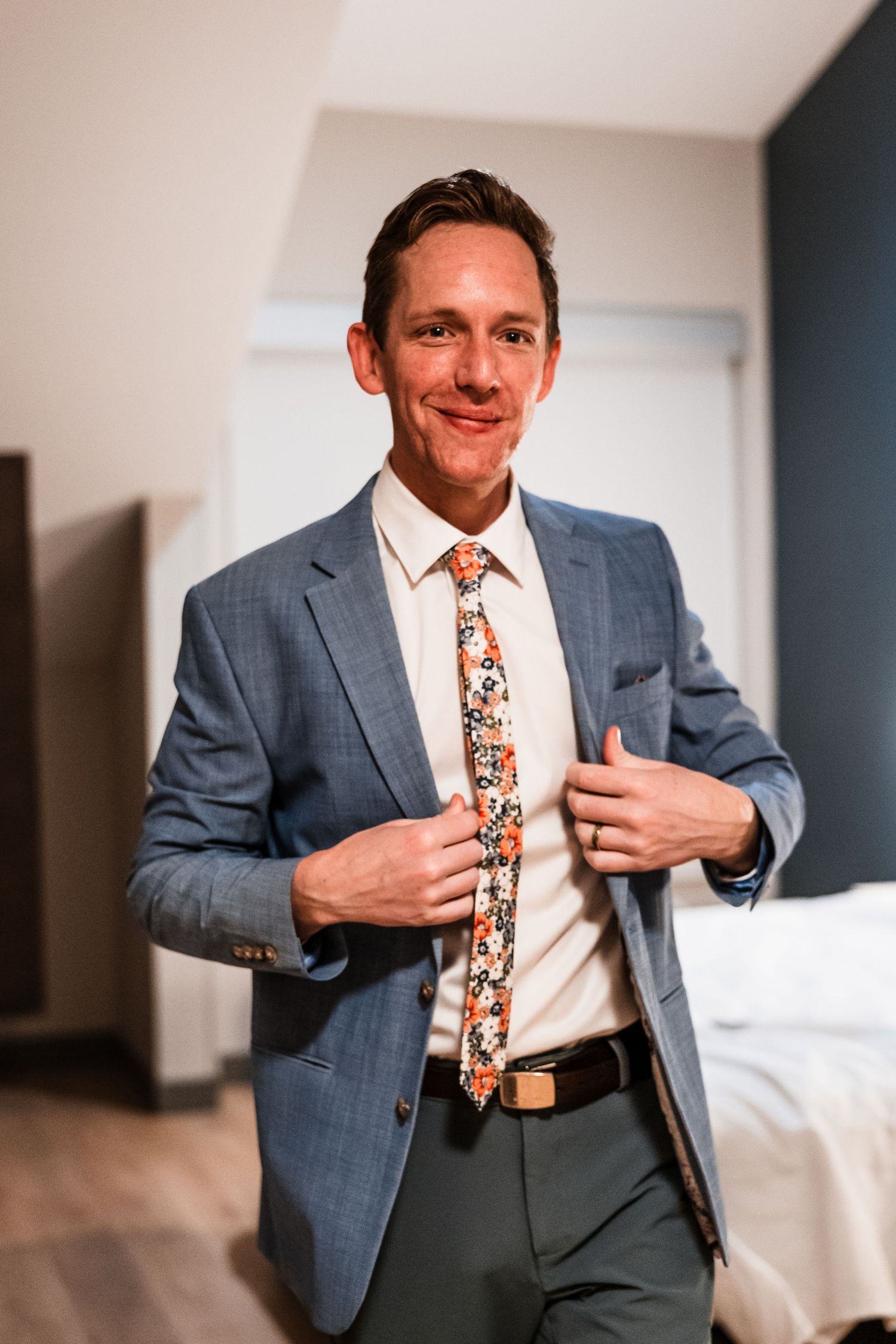 Groom in blue suit and floral tie smiling while adjusting jacket in hotel room before wedding.