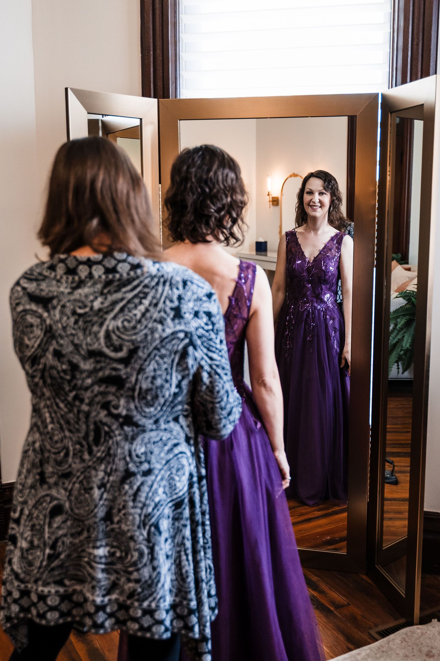 Woman in purple sequin gown smiling at her reflection in a full-length mirror during wedding preparations.