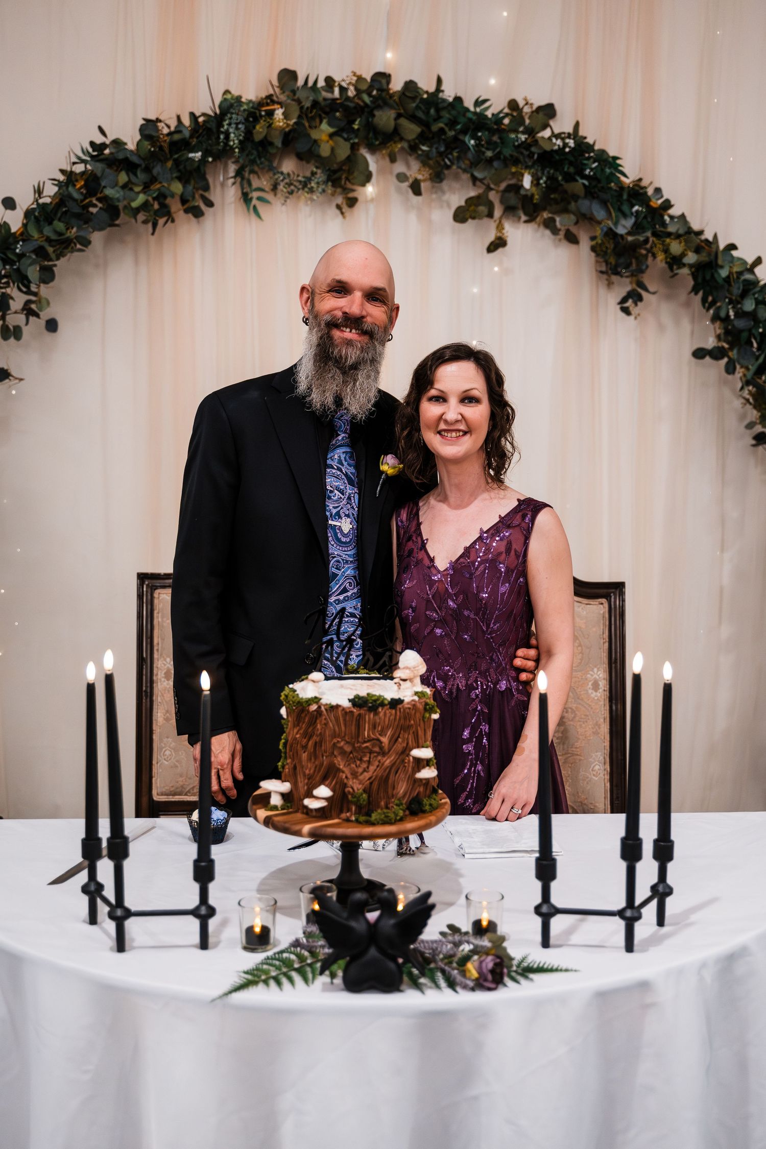 Couple poses at sweetheart table with mushroom wedding cake and black taper candles.