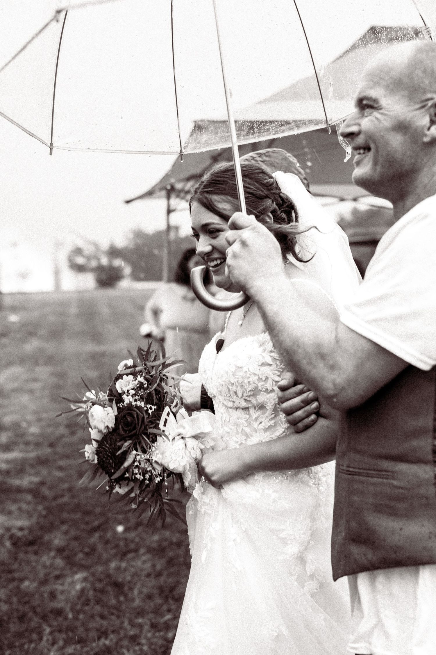 A joyful wedding moment captured in black and white as a couple shares an umbrella while holding a floral bouquet.
