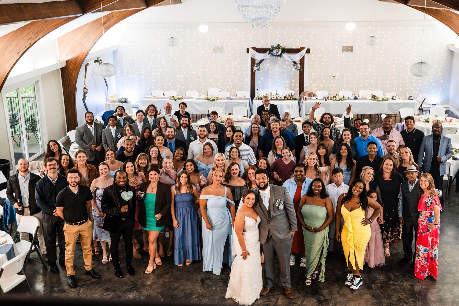 Large group photo of wedding guests gathered under an arch in an elegant indoor venue.