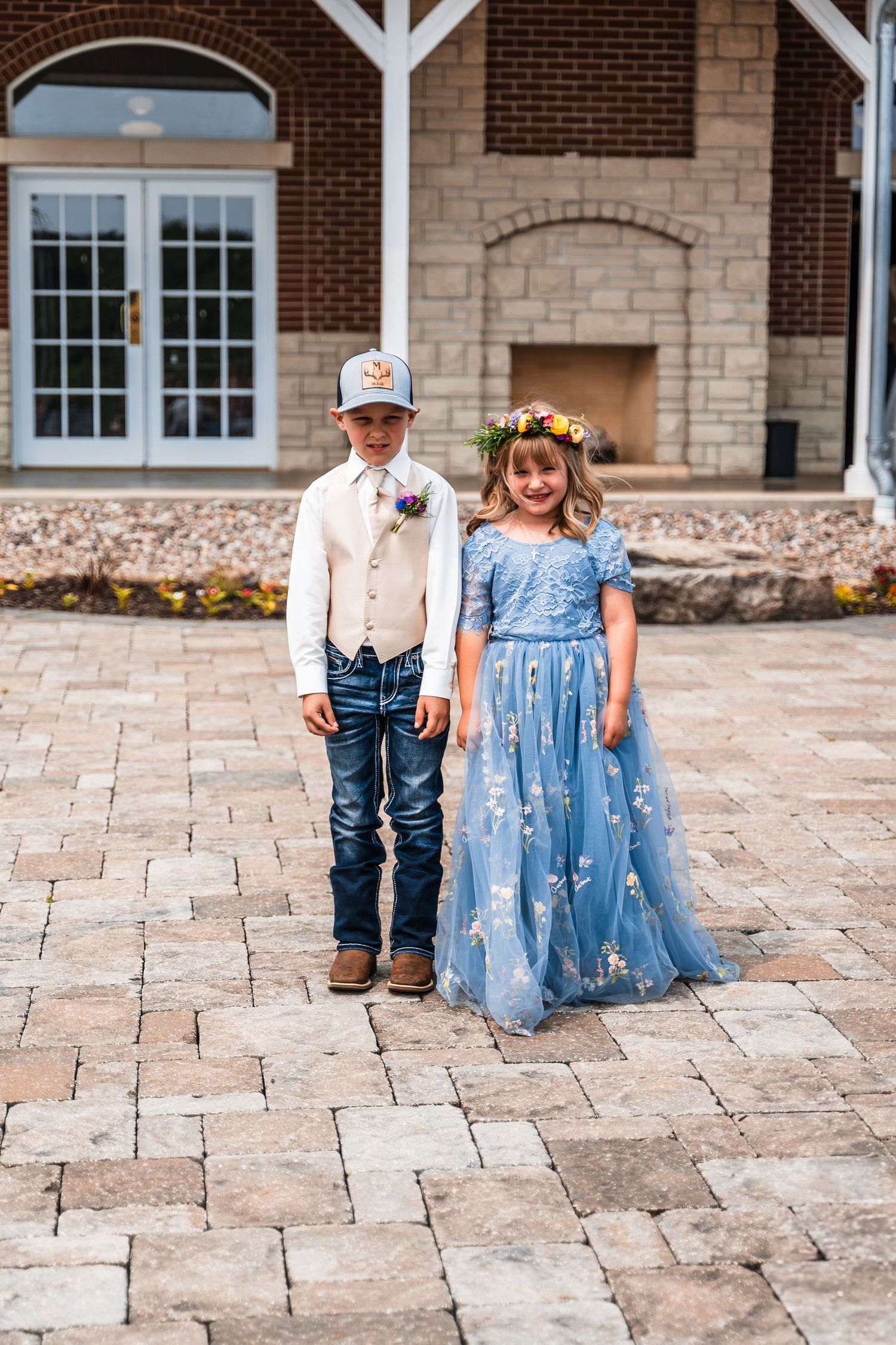 Young attendants in formal wear pose on stone patio outside brick and stone building.