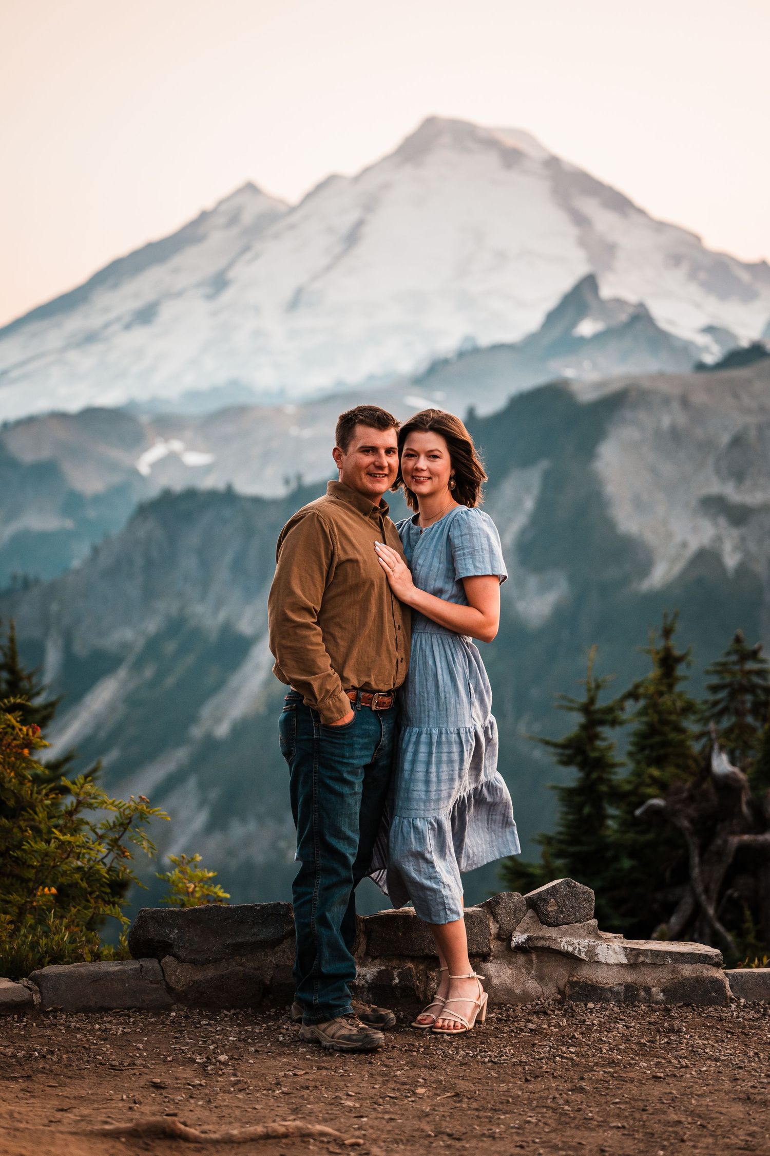 A couple sits together on rocks with a majestic snow-capped mountain peak in the background at sunset.
