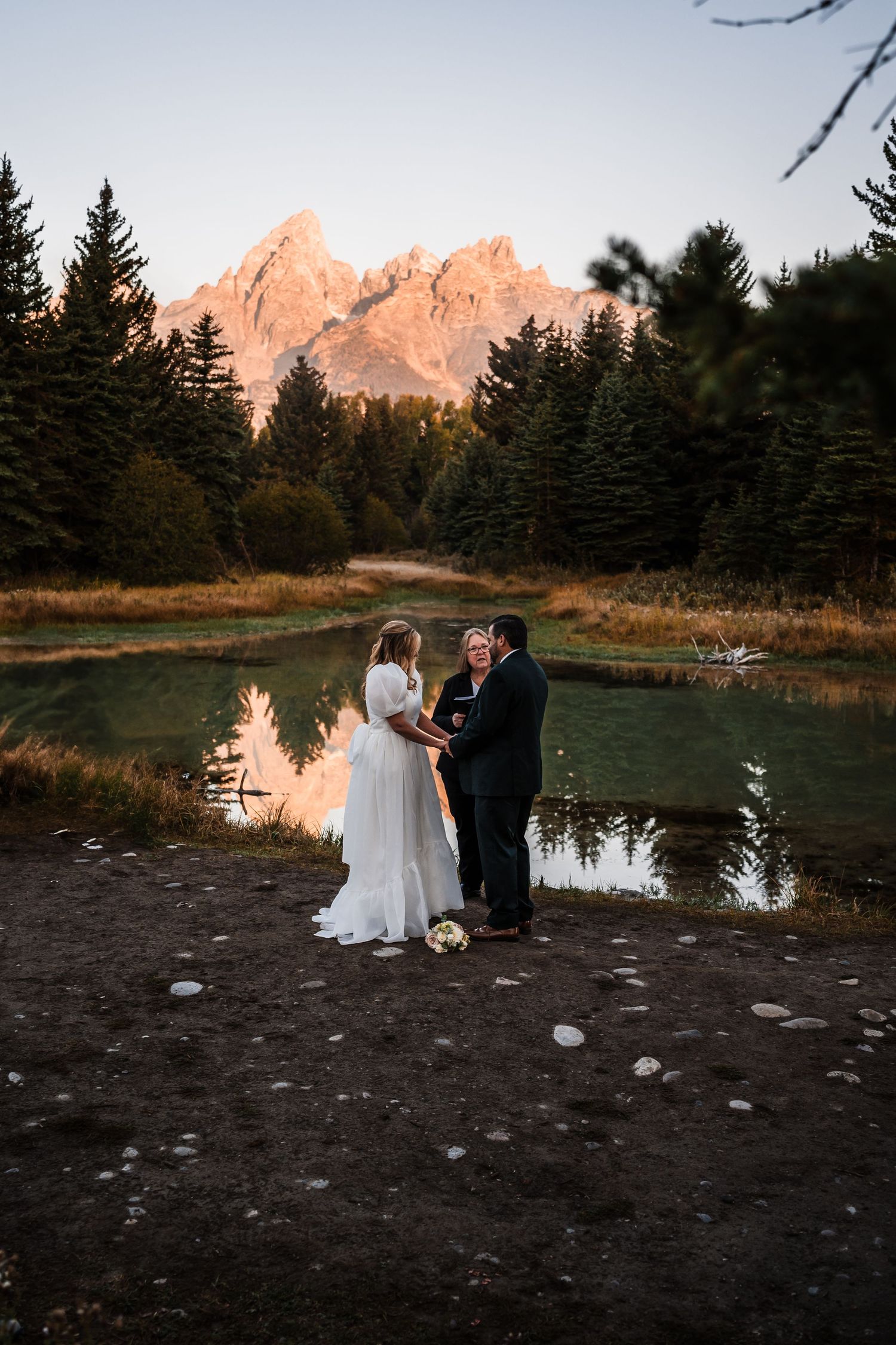 A wedding couple shares an intimate moment at sunset near a mountain lake surrounded by evergreen trees.