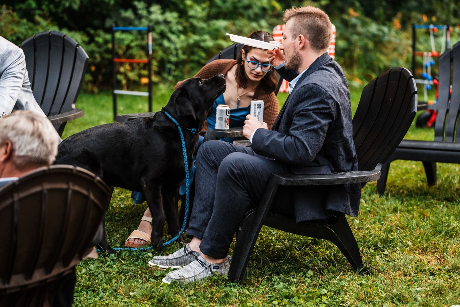A guest enjoys a drink while sitting in an Adirondack chair with a black dog.