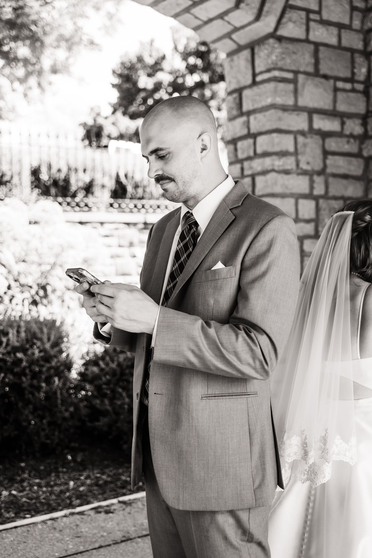 A person in a gray suit checks their phone while standing near a brick wall outdoors.