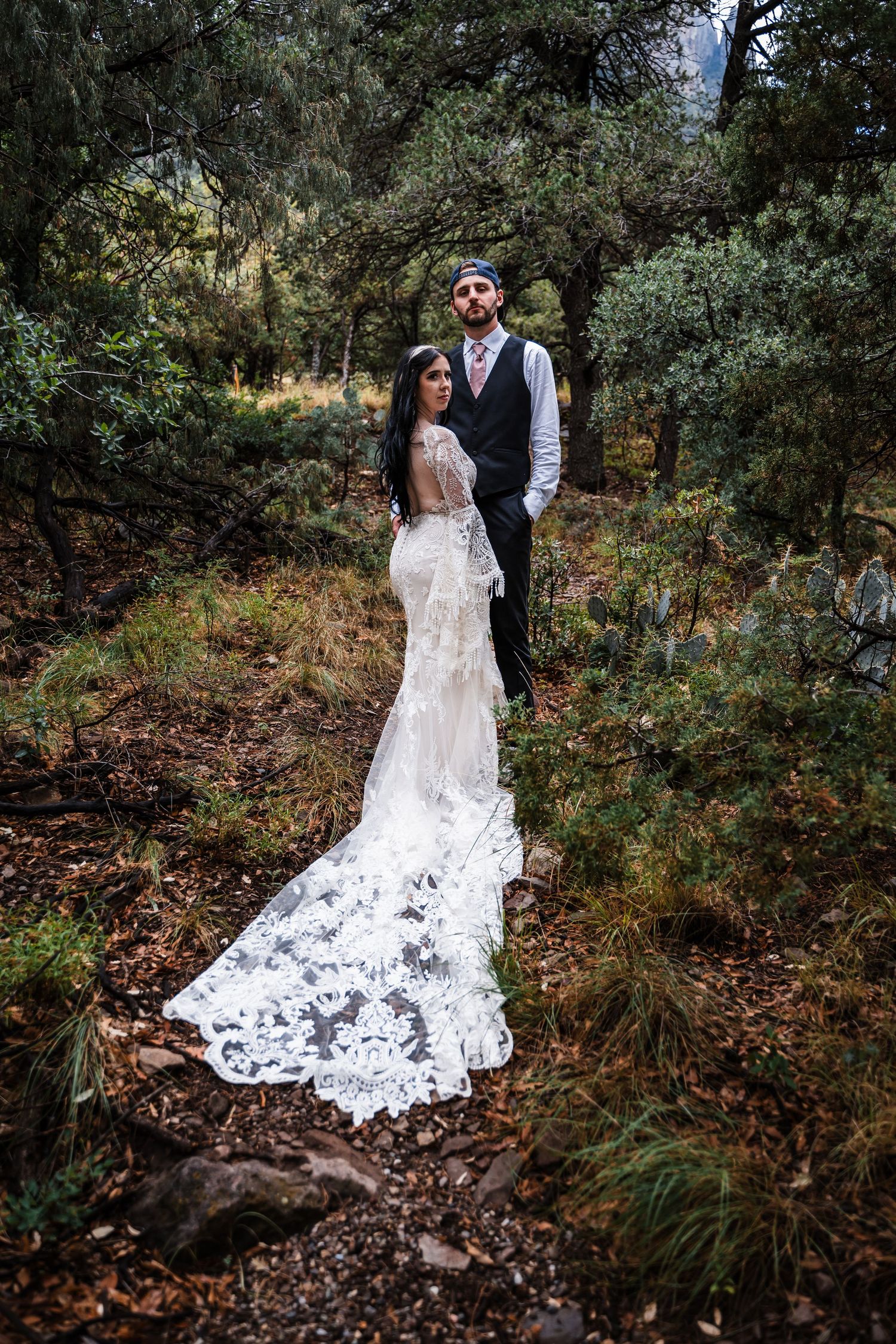A bride in lace gown with long train poses with partner in a wooded forest setting.