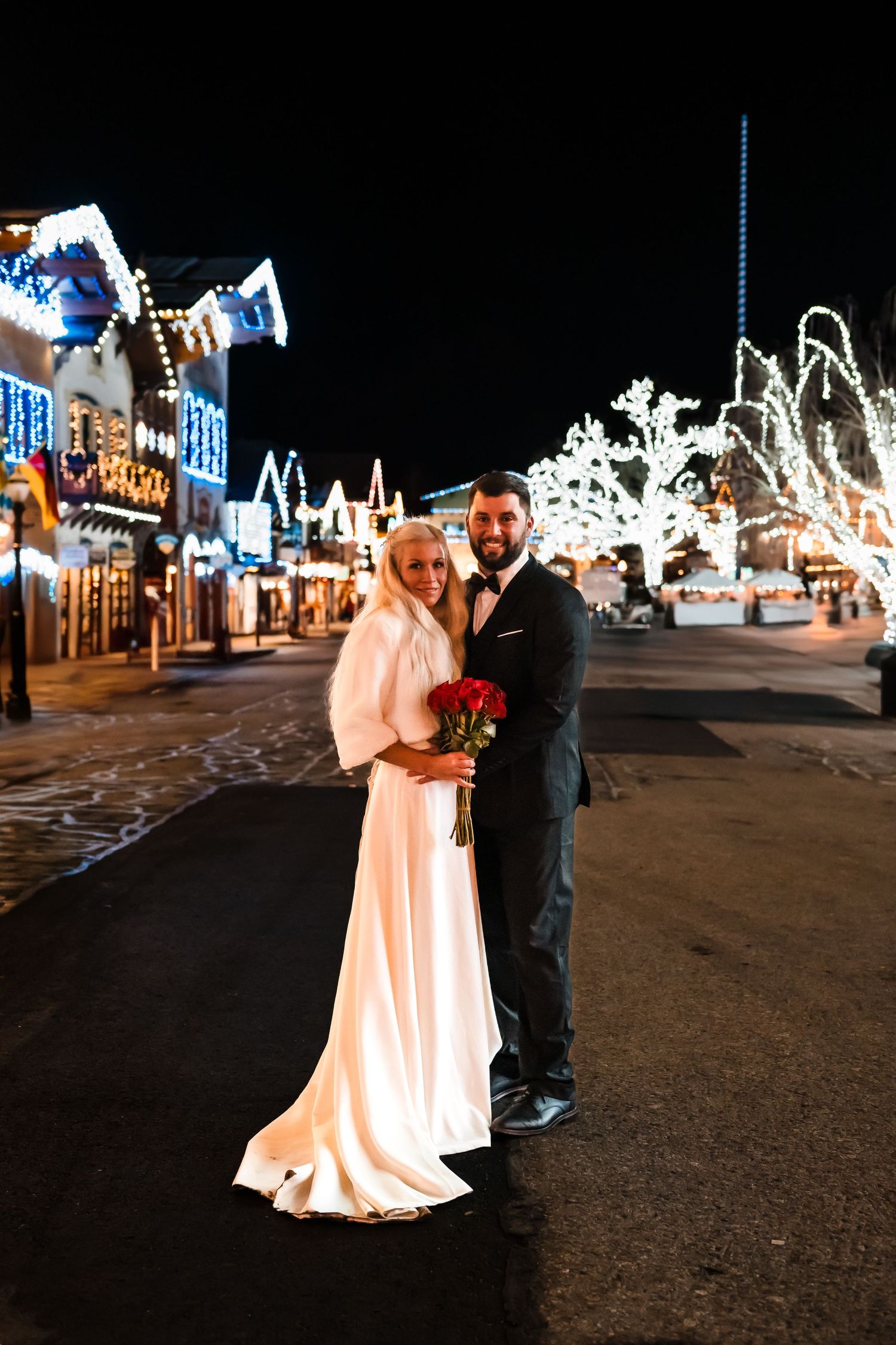 A winter wedding photo on a festive street with holiday lights and decorations.