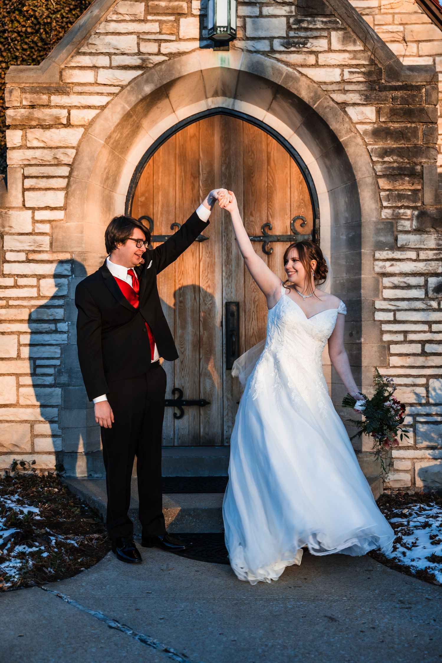 Couple dances and twirls in front of rustic wooden church door surrounded by stone architecture.