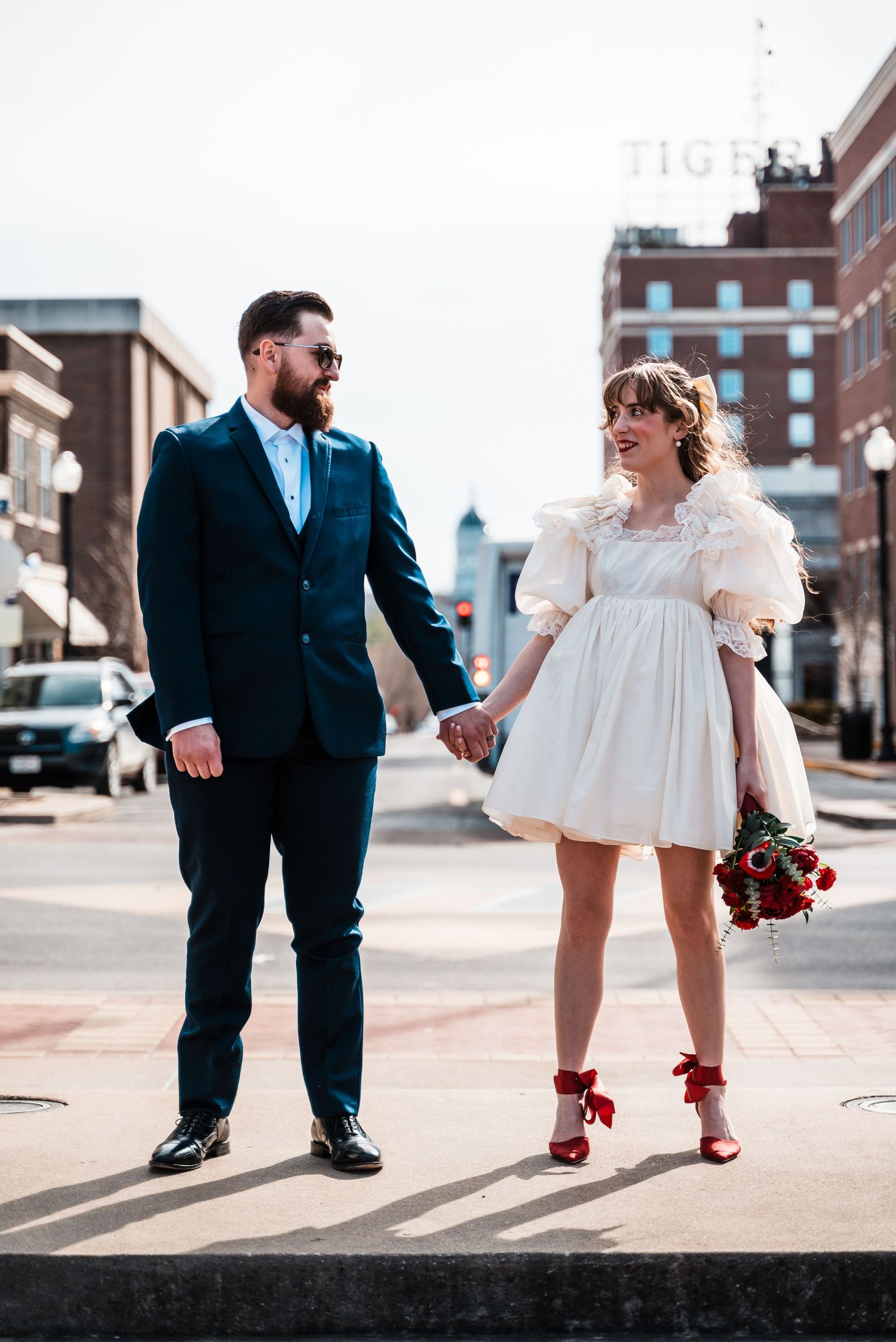 A couple in formal attire poses on a city sidewalk with red roses and matching shoes.