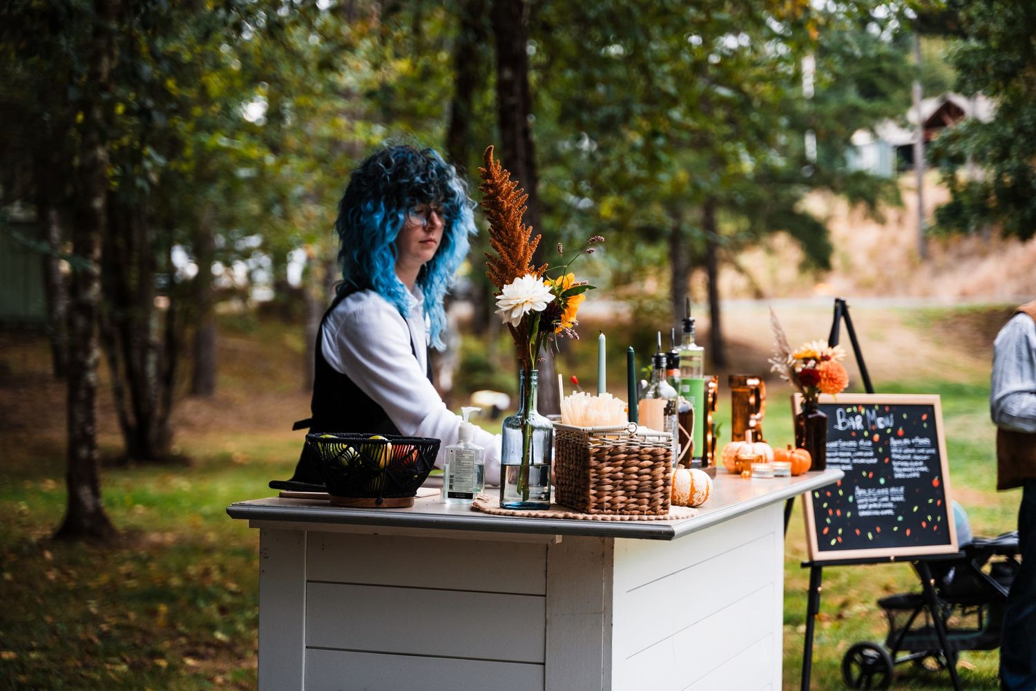 A bartender in blue hair mixes drinks at an outdoor bar station with bottles and glassware.