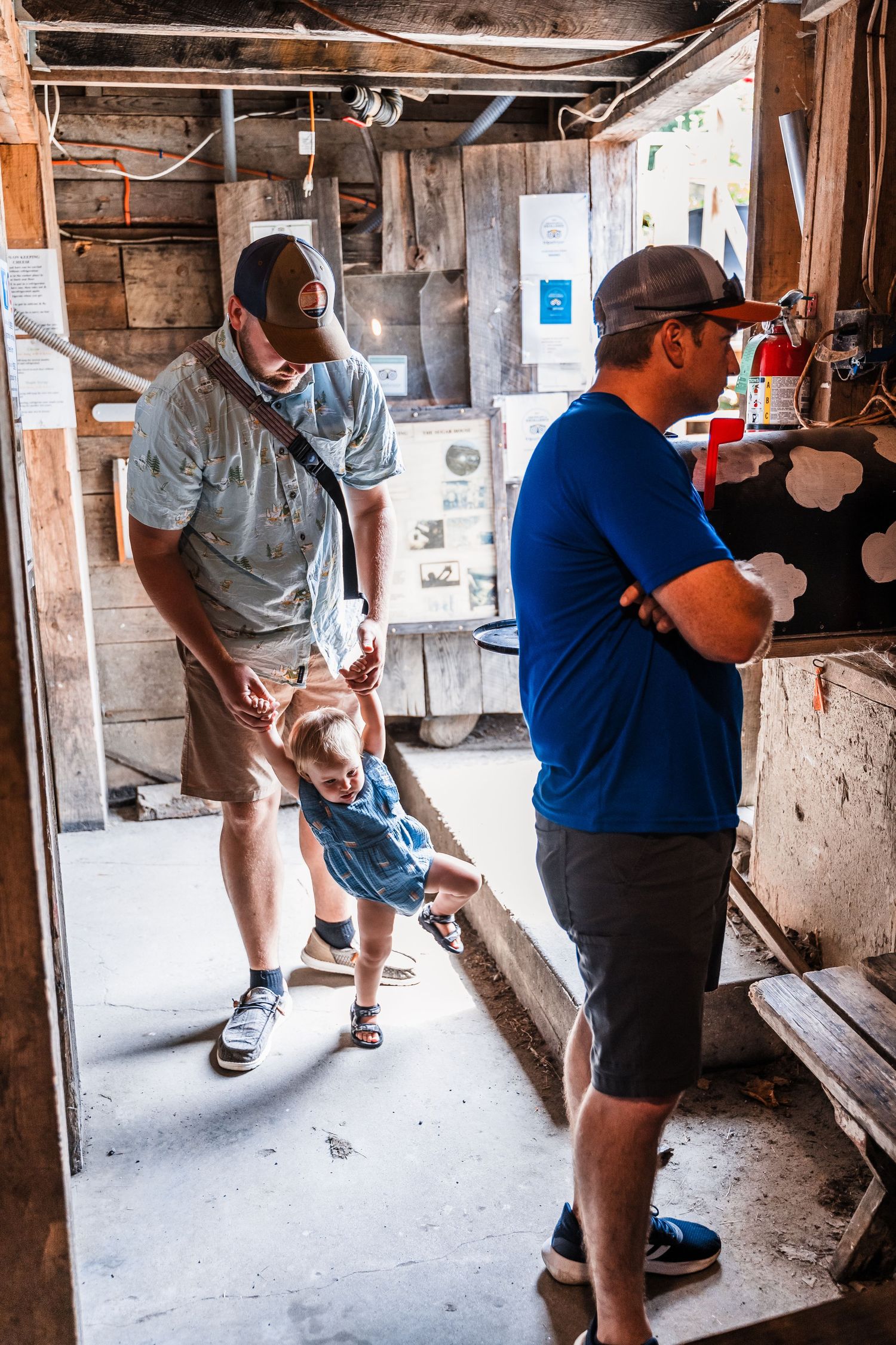 Two adults and a small child stand in a rustic workshop-like space with exposed wooden walls and various tools.