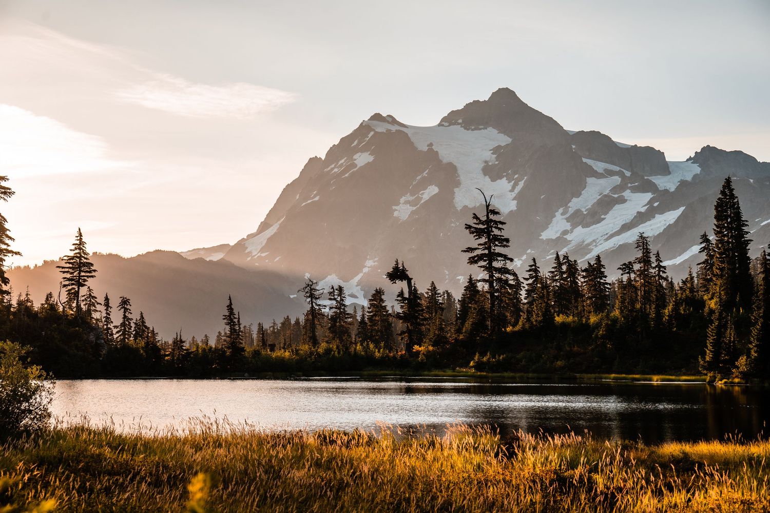Majestic snow-capped mountain peak reflects in serene lake surrounded by evergreen forest at golden sunset.
