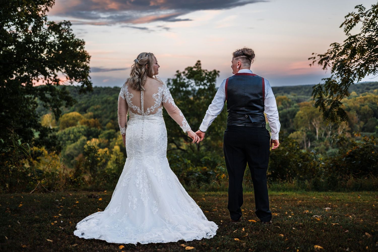 A couple holds hands while viewing a sunset from a hilltop overlook in formal wedding attire.