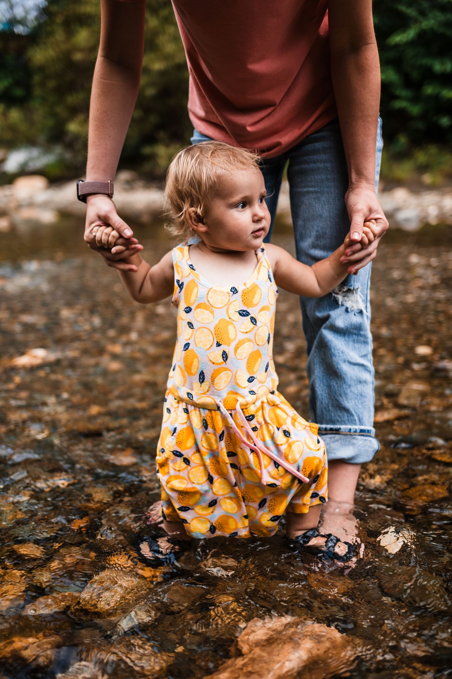 Child in yellow floral dress holding hands while standing in a shallow stream.