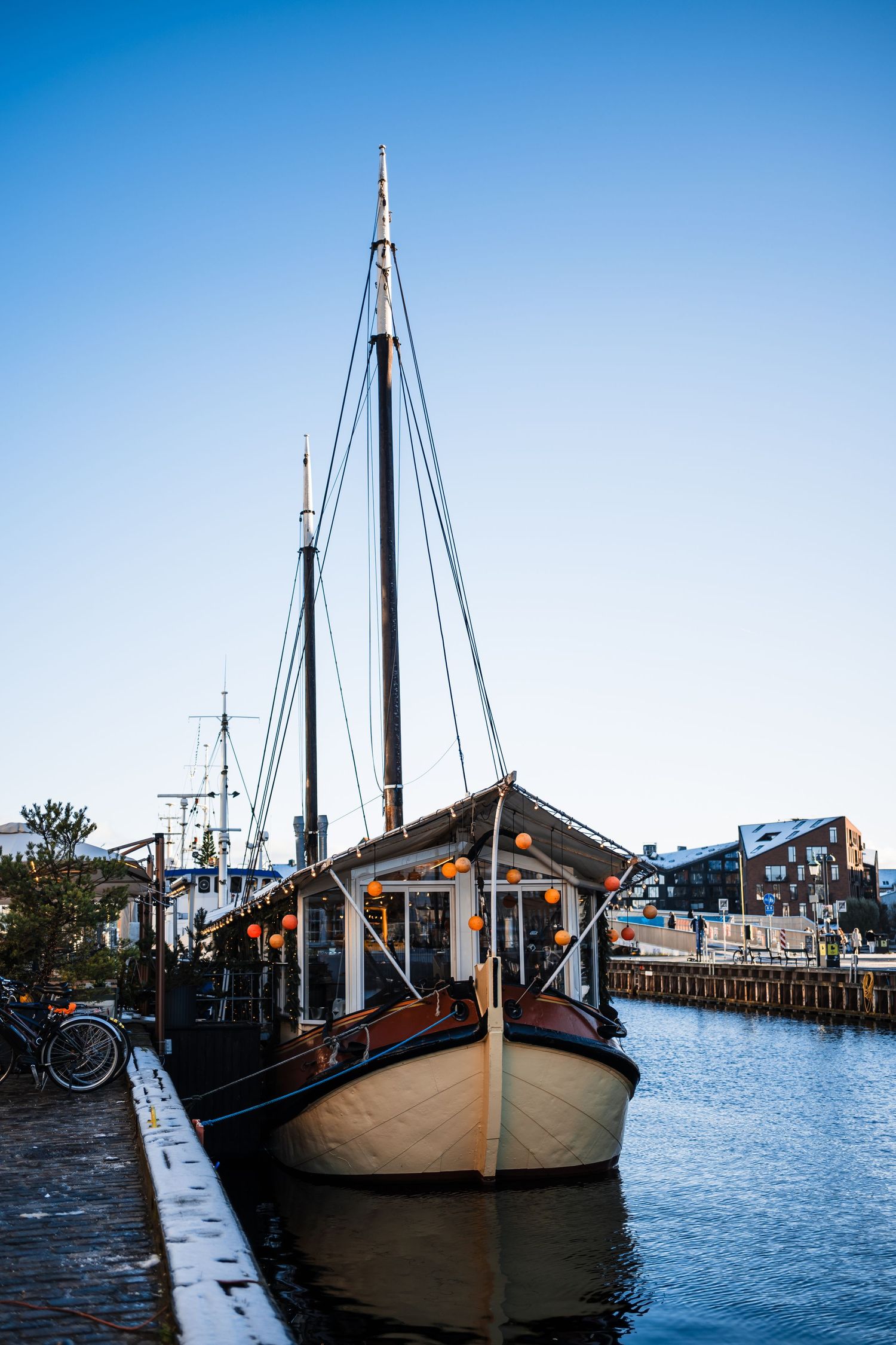 Wooden sailboat with tall masts moored at a marina dock against a bright blue sky.