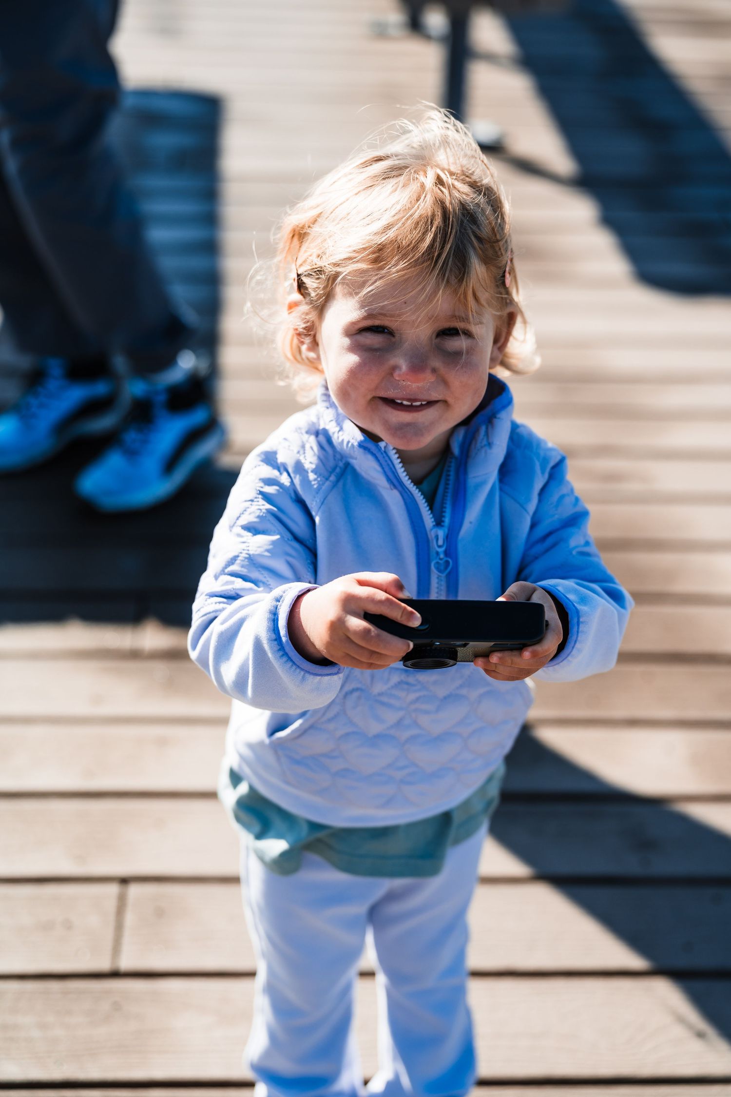 Young child in light blue jacket smiling while holding a device on wooden boardwalk.