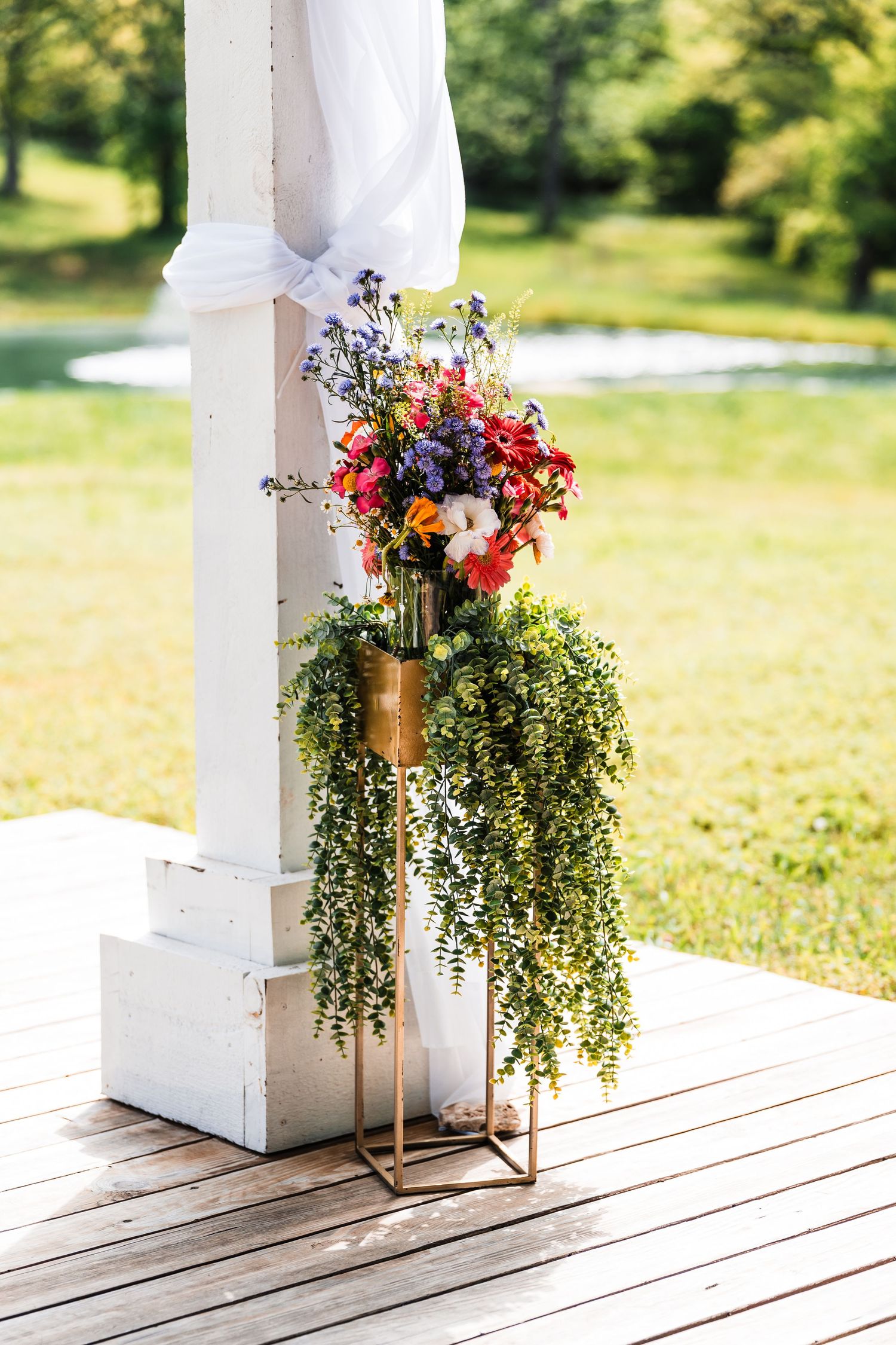 Gold geometric stand with colorful wildflower bouquet and cascading greenery beside white wedding arch outdoors.