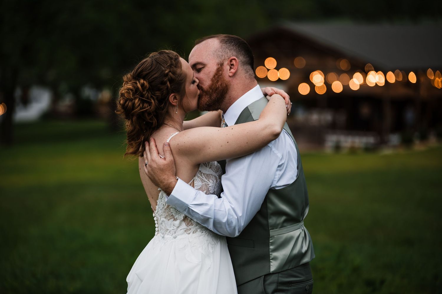 Bride and groom share romantic outdoor kiss at dusk with string lights behind them.