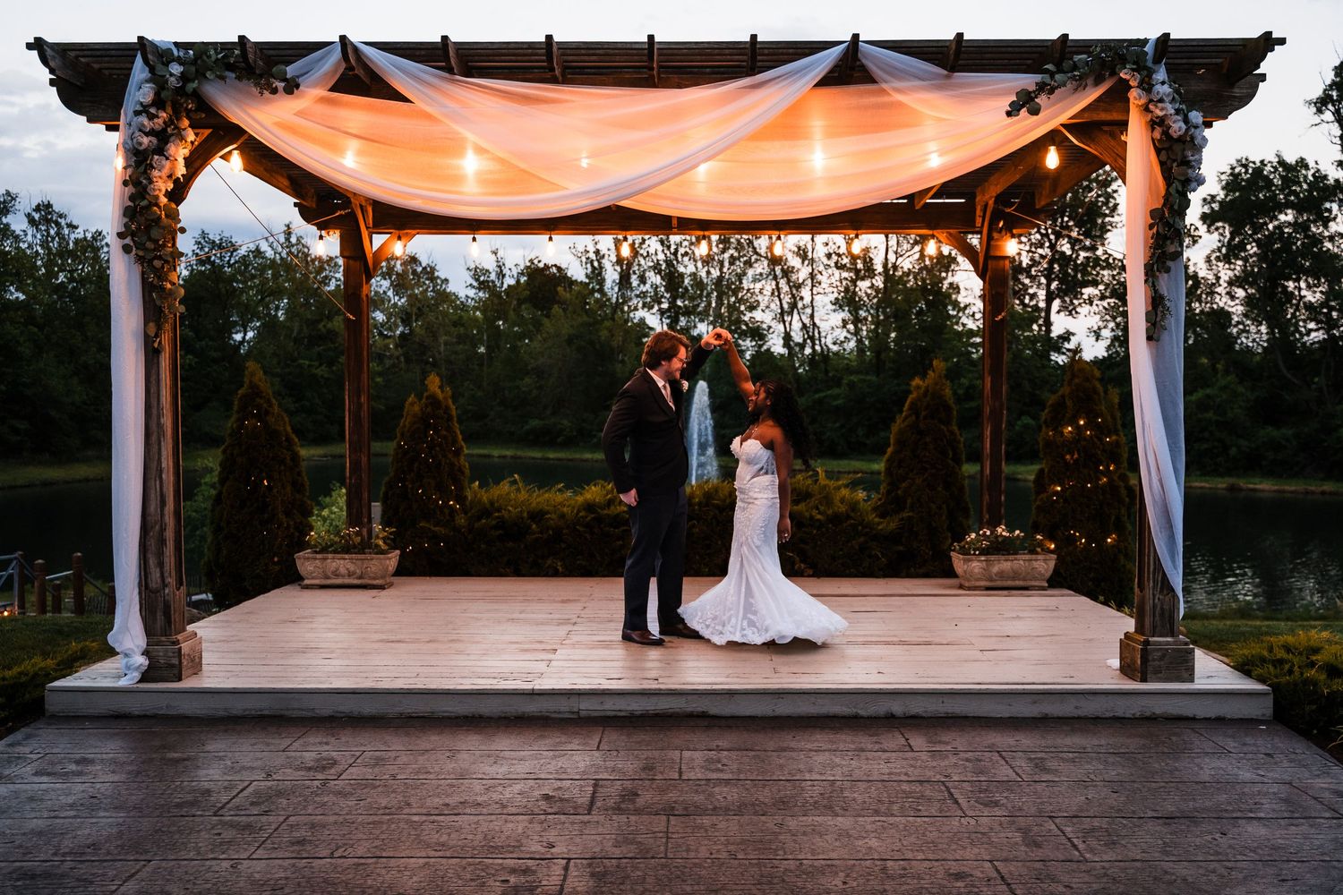 Bride and groom share a romantic moment under a draped pergola with string lights at dusk near a serene pond.