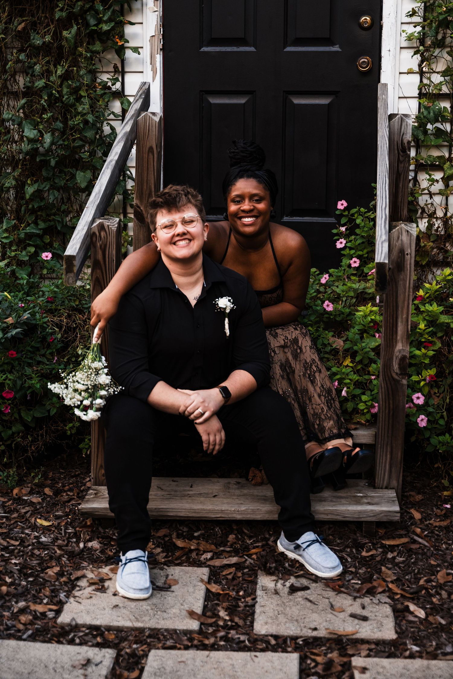 A couple sits together on wooden steps in front of a black door surrounded by flowering plants.