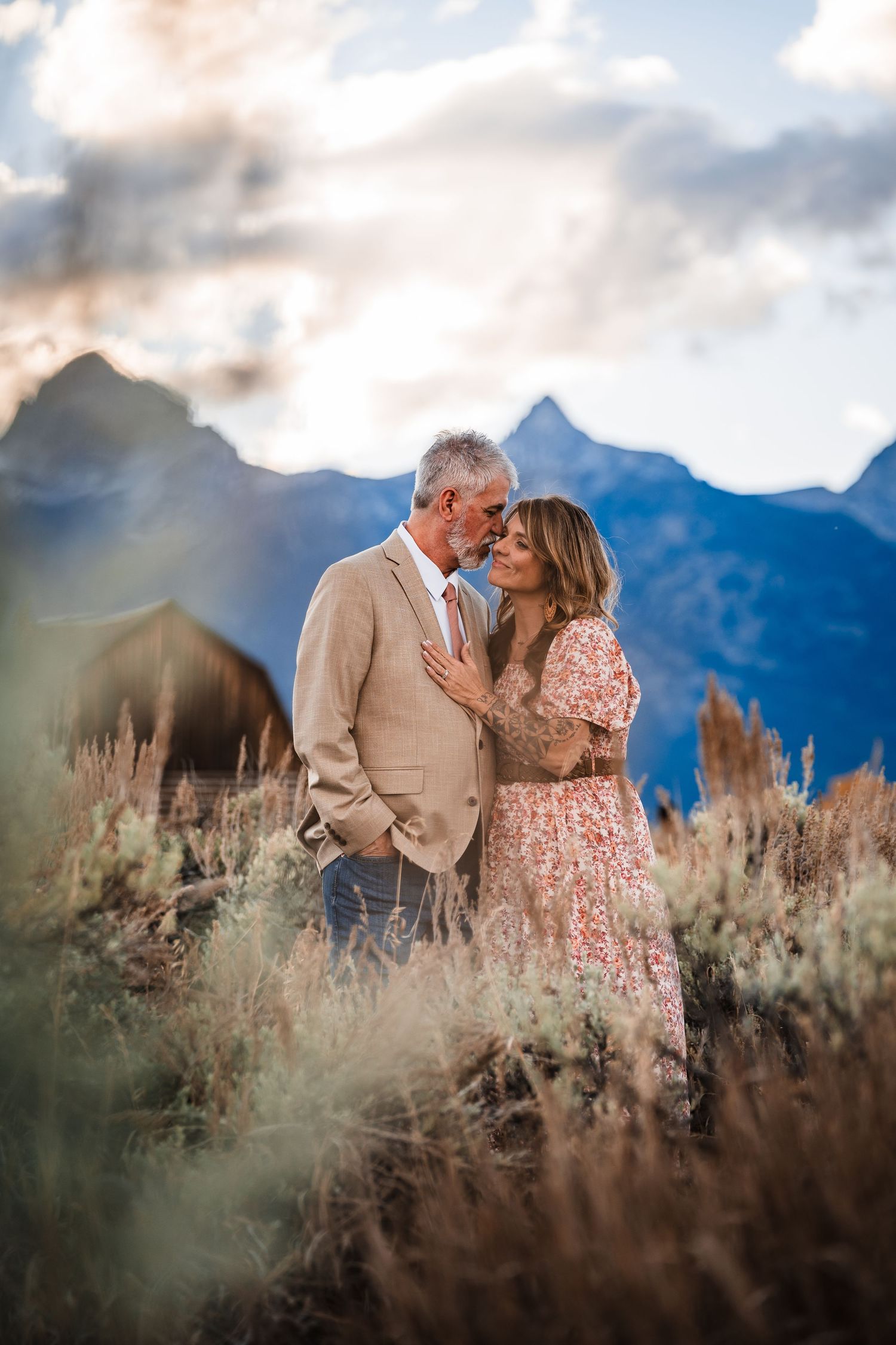 Couple shares romantic moments in a field of tall grass with majestic mountain peaks in the background at sunset.