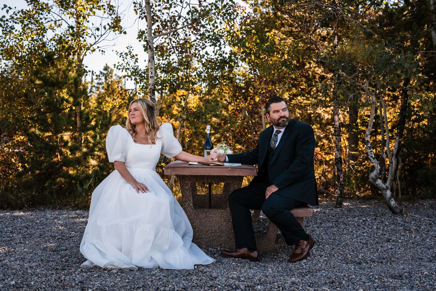Newlywed couple sits together on rustic bench during sunset golden hour photo session in outdoor setting.