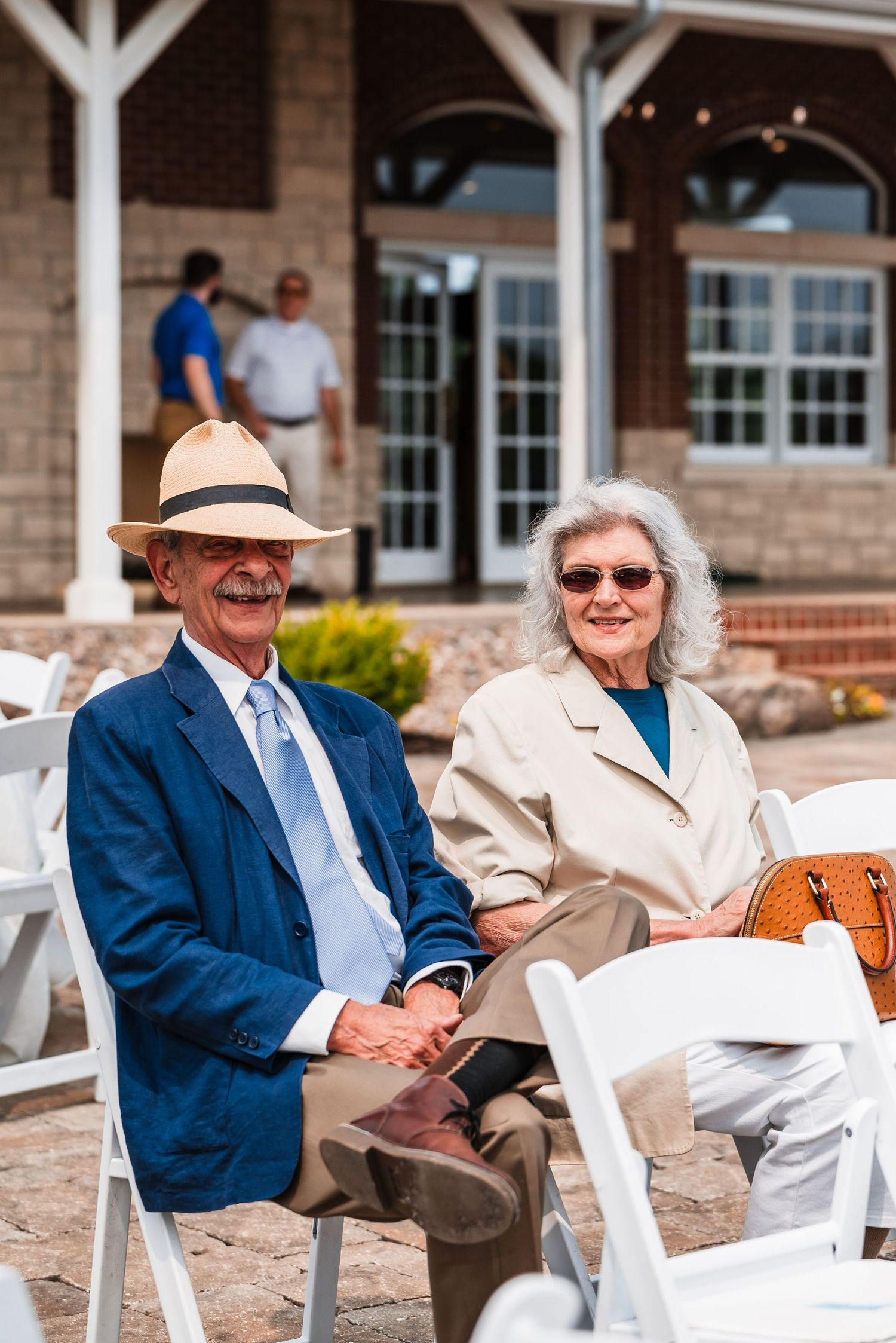 Wedding guests in formal attire sitting outdoors on white chairs at a brick venue.