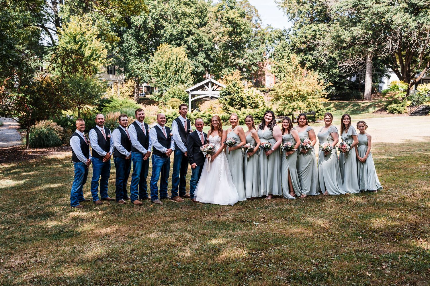 Wedding party members in coordinating blue and gray attire pose together on a sunny grassy lawn.