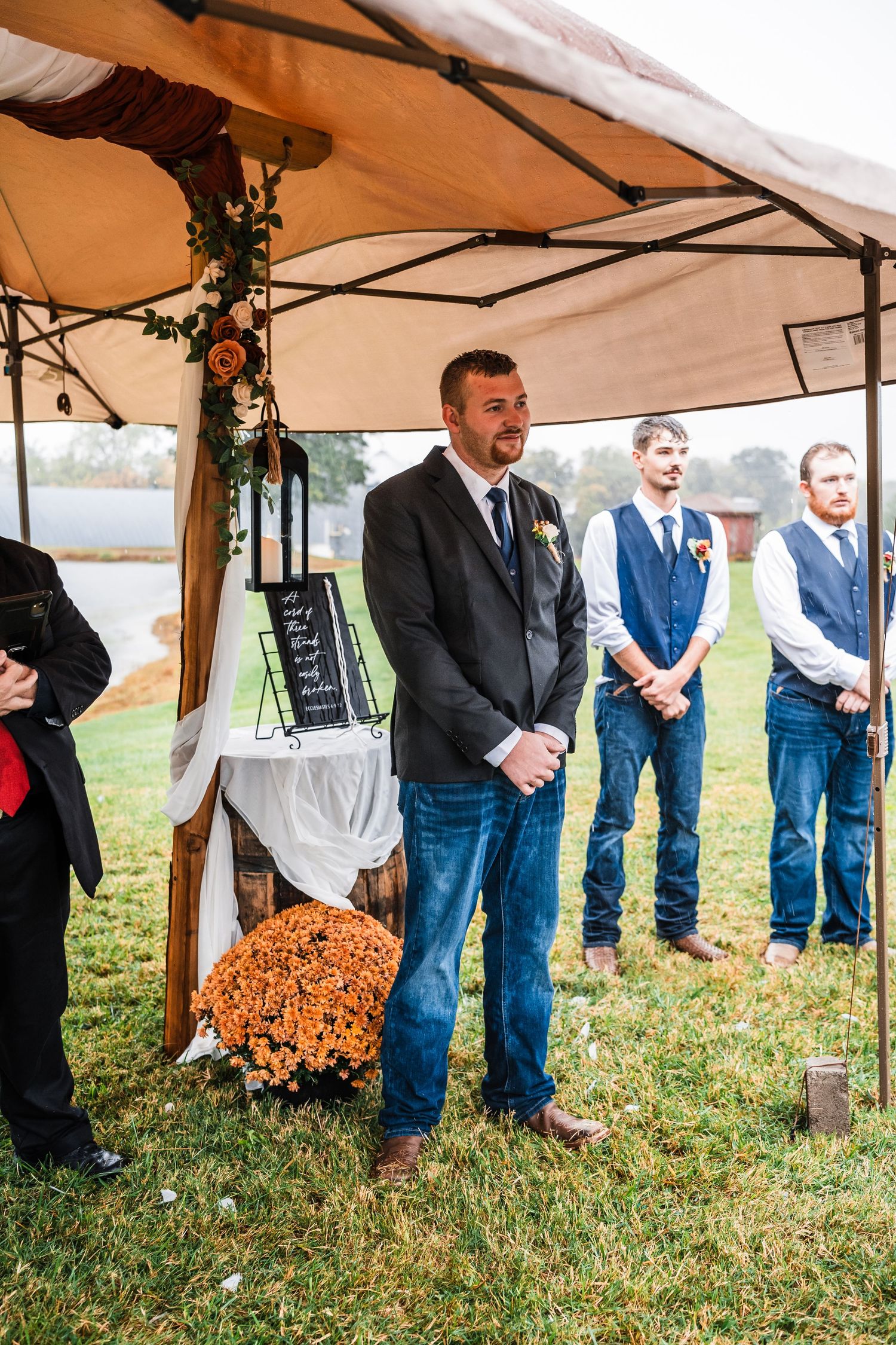 Wedding party members stand under a tent decorated with greenery and orange flowers during an outdoor ceremony.